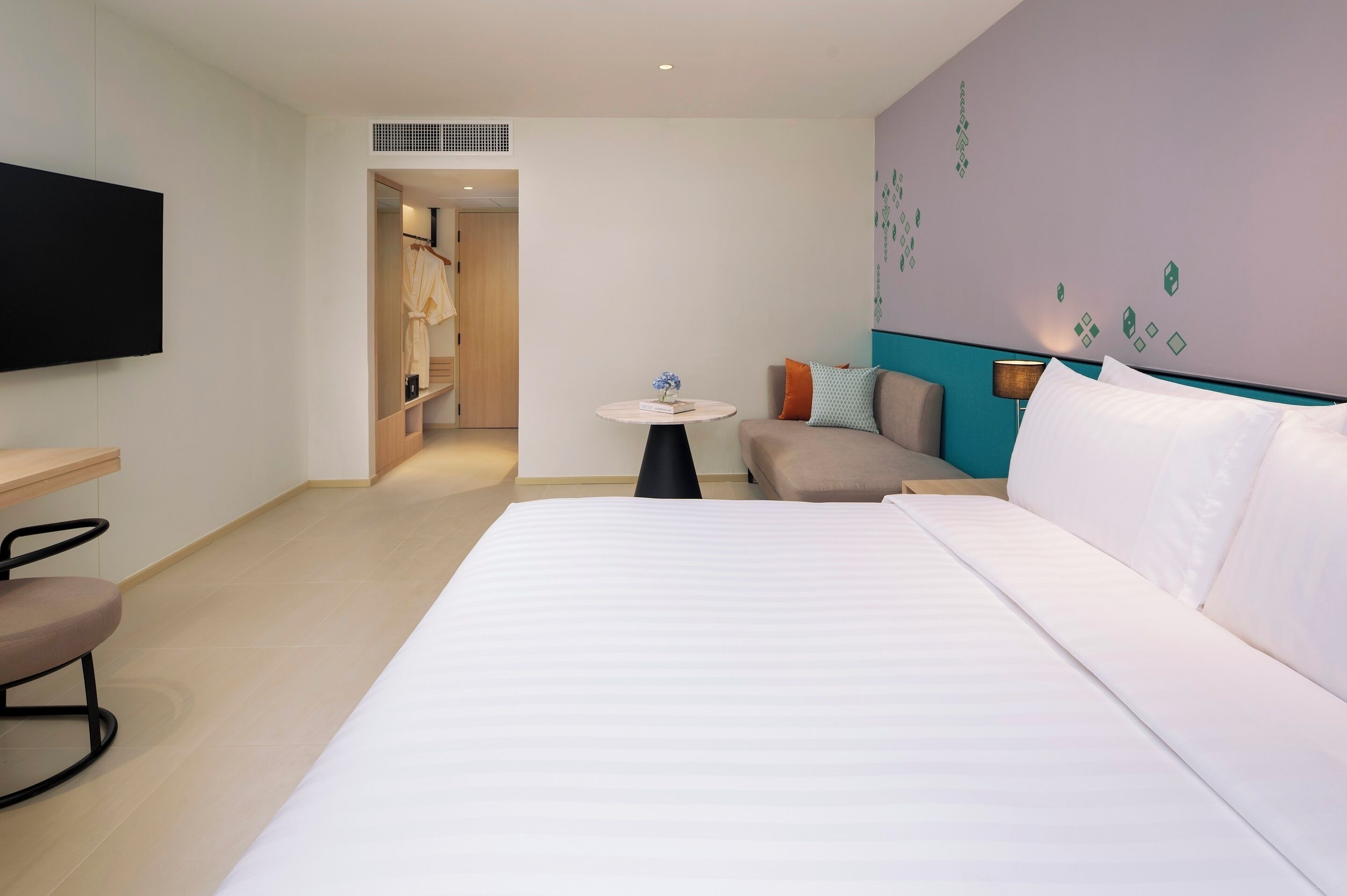 A hotel room with a bed, a TV, a small table with a plant, a sofa with cushions, and a wardrobe.