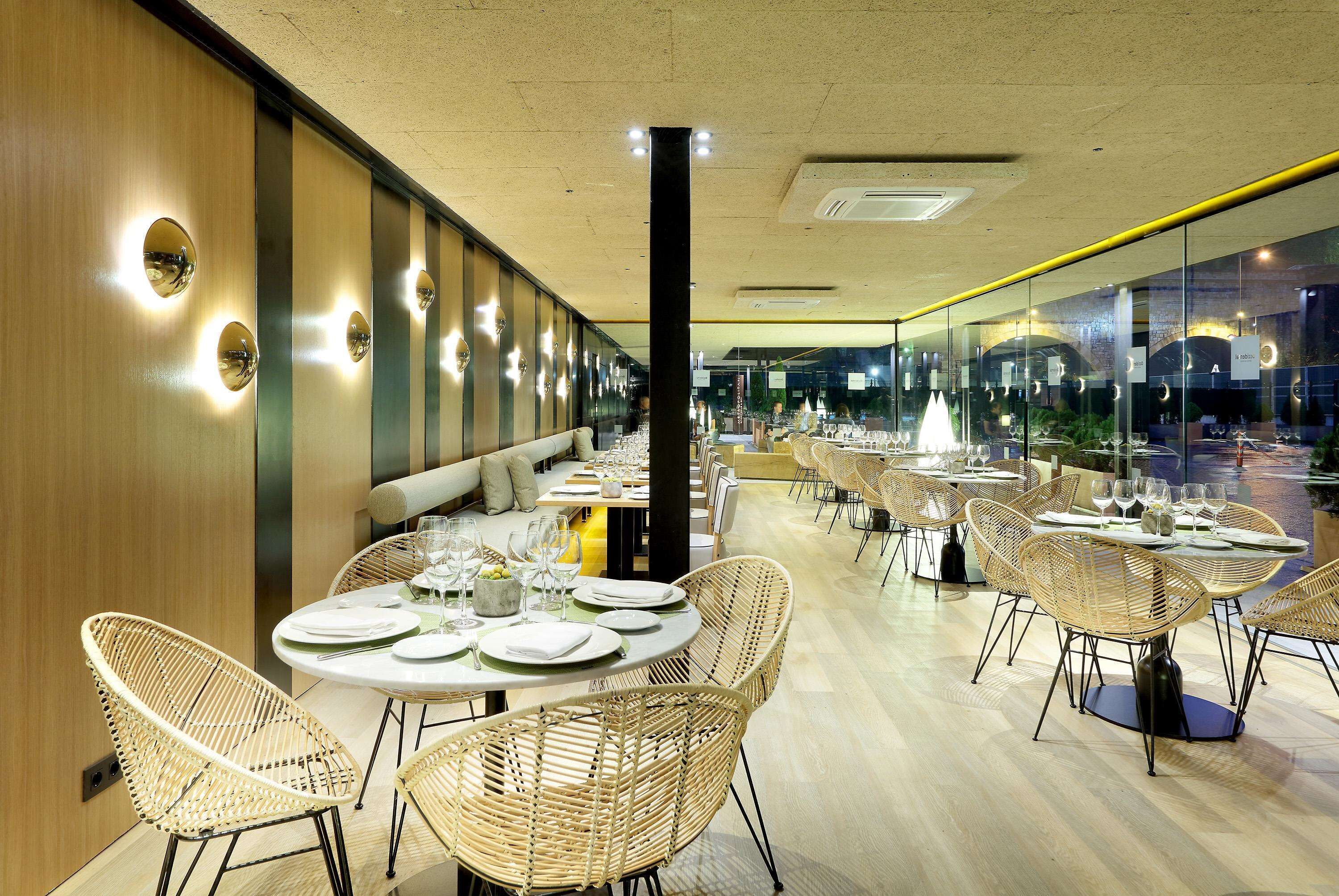 Un restaurant moderne avec des tables rondes et des chaises en osier, un mur en bois orné de luminaires circulaires et une vue dégagée sur l’extérieur grâce à de grandes fenêtres.