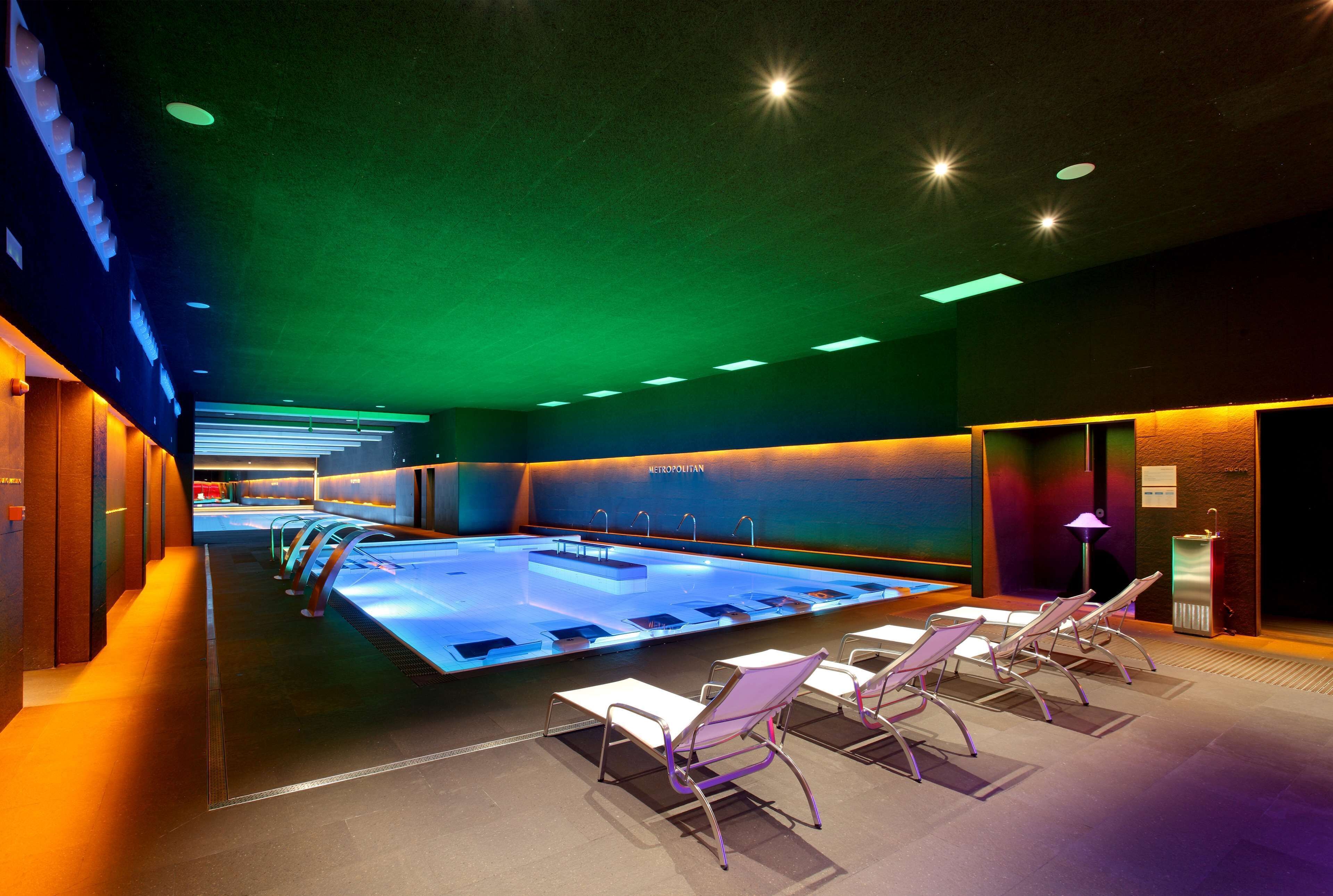 Une piscine intérieure moderne avec un plafond éclairé de lumière verte, plusieurs couloirs de nage et des chaises longues.