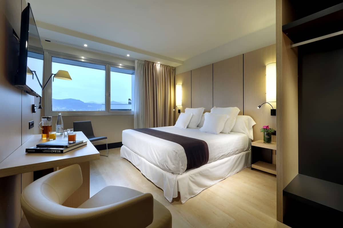 A modern hotel room with a large bed, a desk with a laptop, a chair, and a TV mounted on the wall.