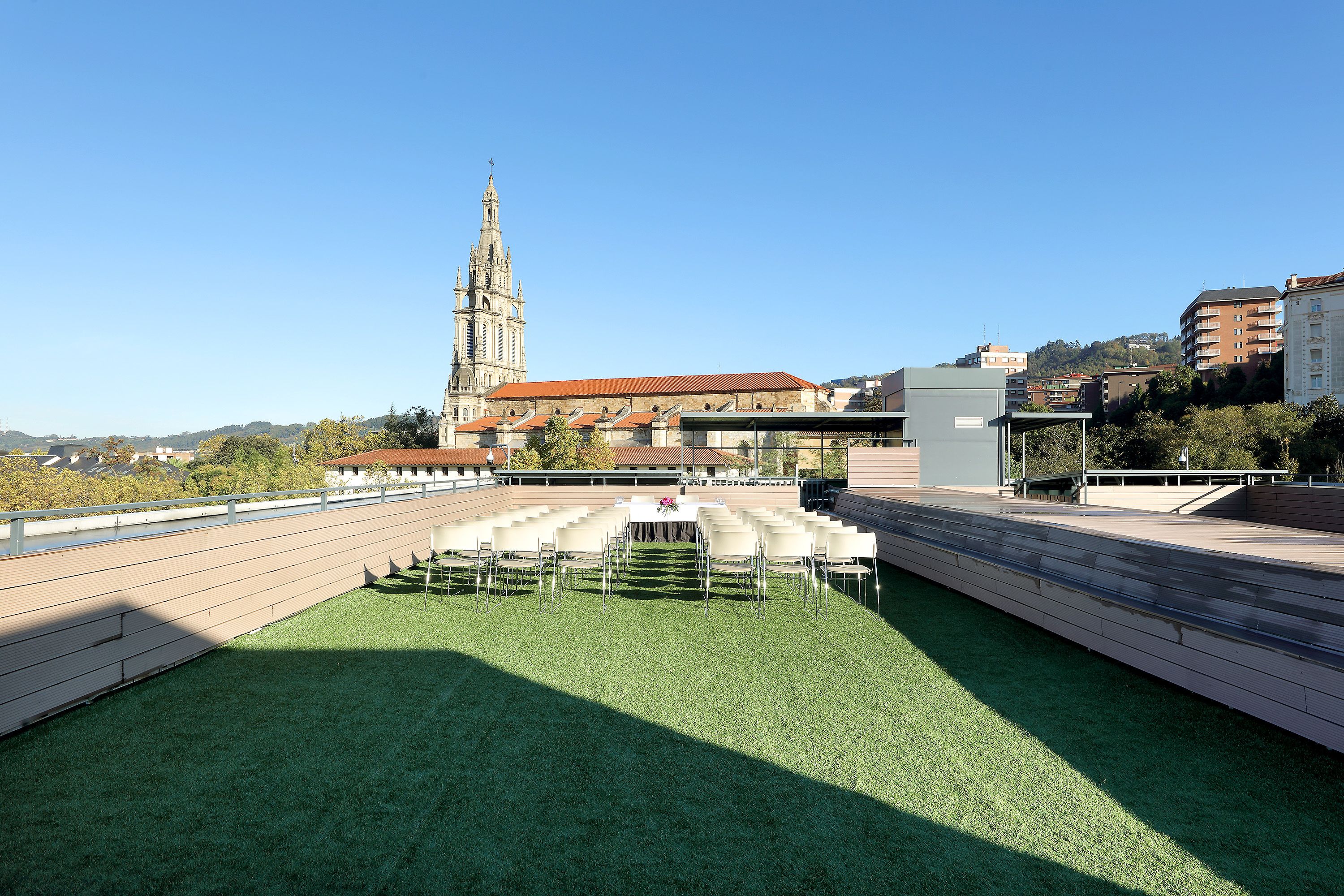 A rooftop terrace with a green lawn, white chairs, and a table with a vase of flowers. In the background, there is a church with a tall tower and a building with a red roof.
