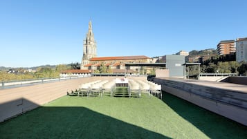 Una terrazza sul tetto con un prato verde, sedie bianche e un tavolo con un vaso di fiori. Sullo sfondo, una chiesa con un'alta torre e un edificio dal tetto rosso.