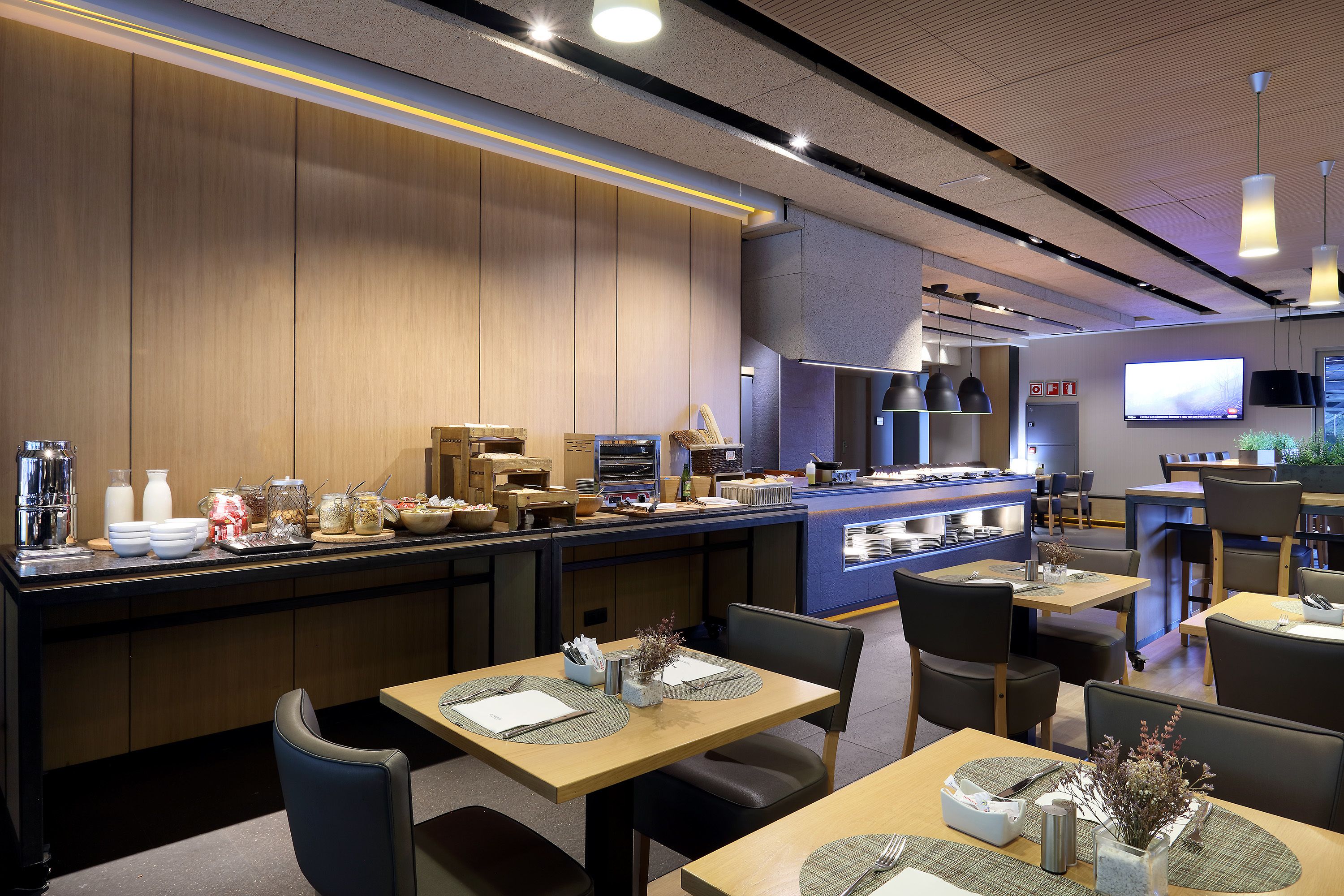 Ein modernes Restaurant mit Holzwandverkleidung, einer Theke mit verschiedenen Speisen und mehreren Tischen mit Stühlen.