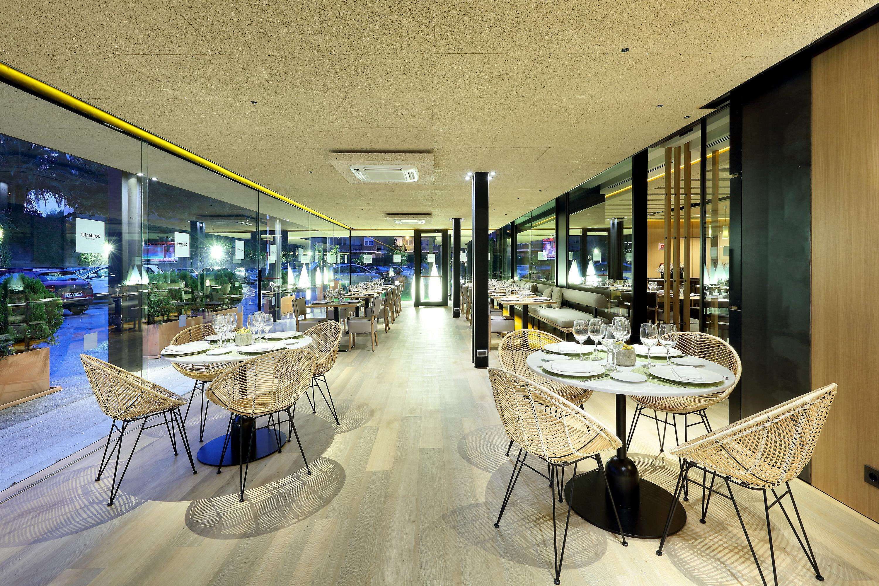 A modern restaurant with round tables and wicker chairs, large windows offering a view of the street, and a clean, contemporary interior design.