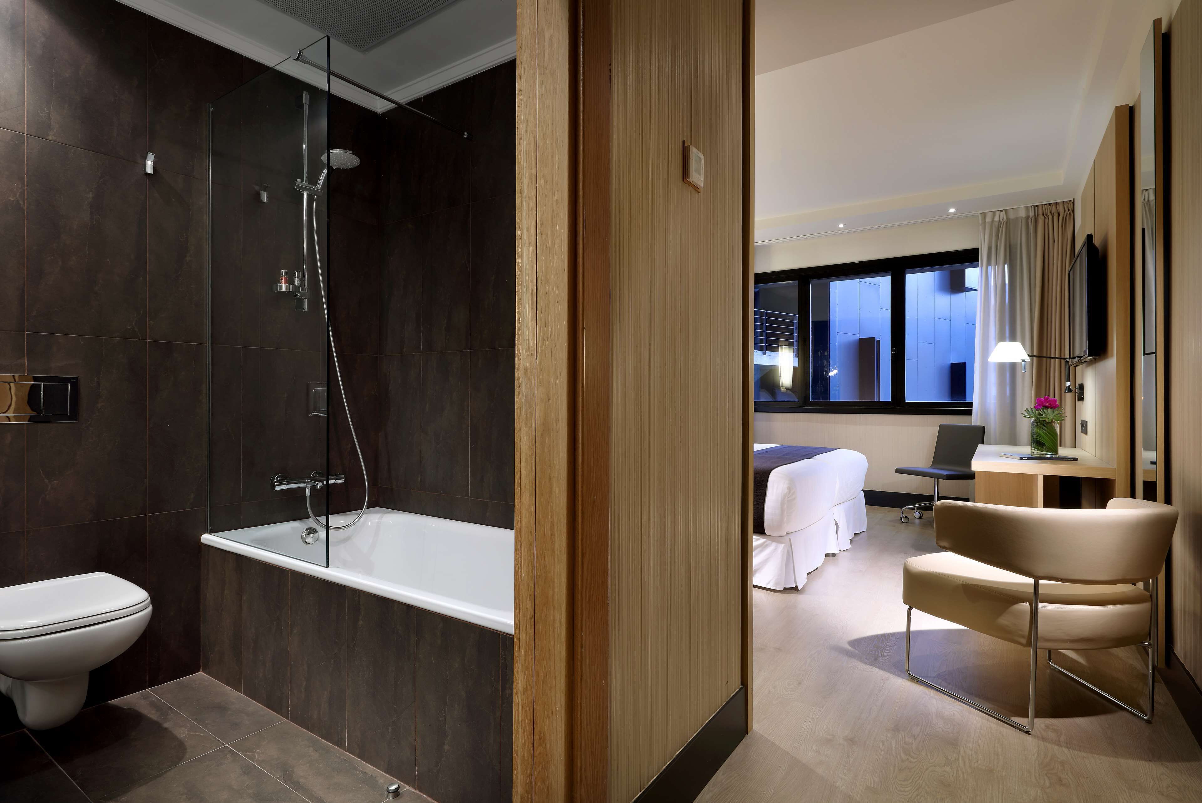 Un bagno moderno con doccia vetro, vasca e WC. Nella stanza adiacente, un letto con scrivania e sedia.