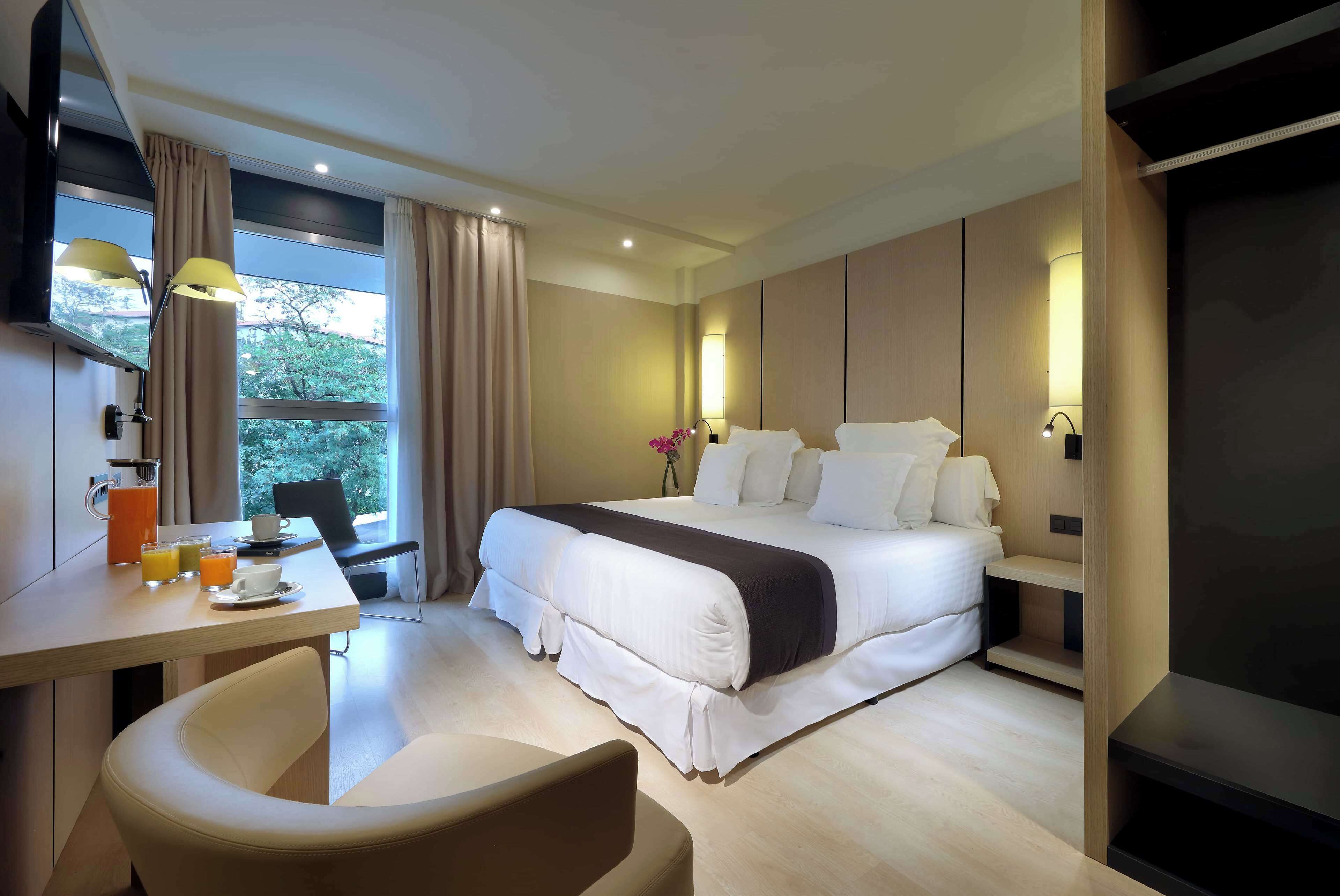 A modern hotel room with a large bed, a desk with a chair, a TV, and a window with curtains.