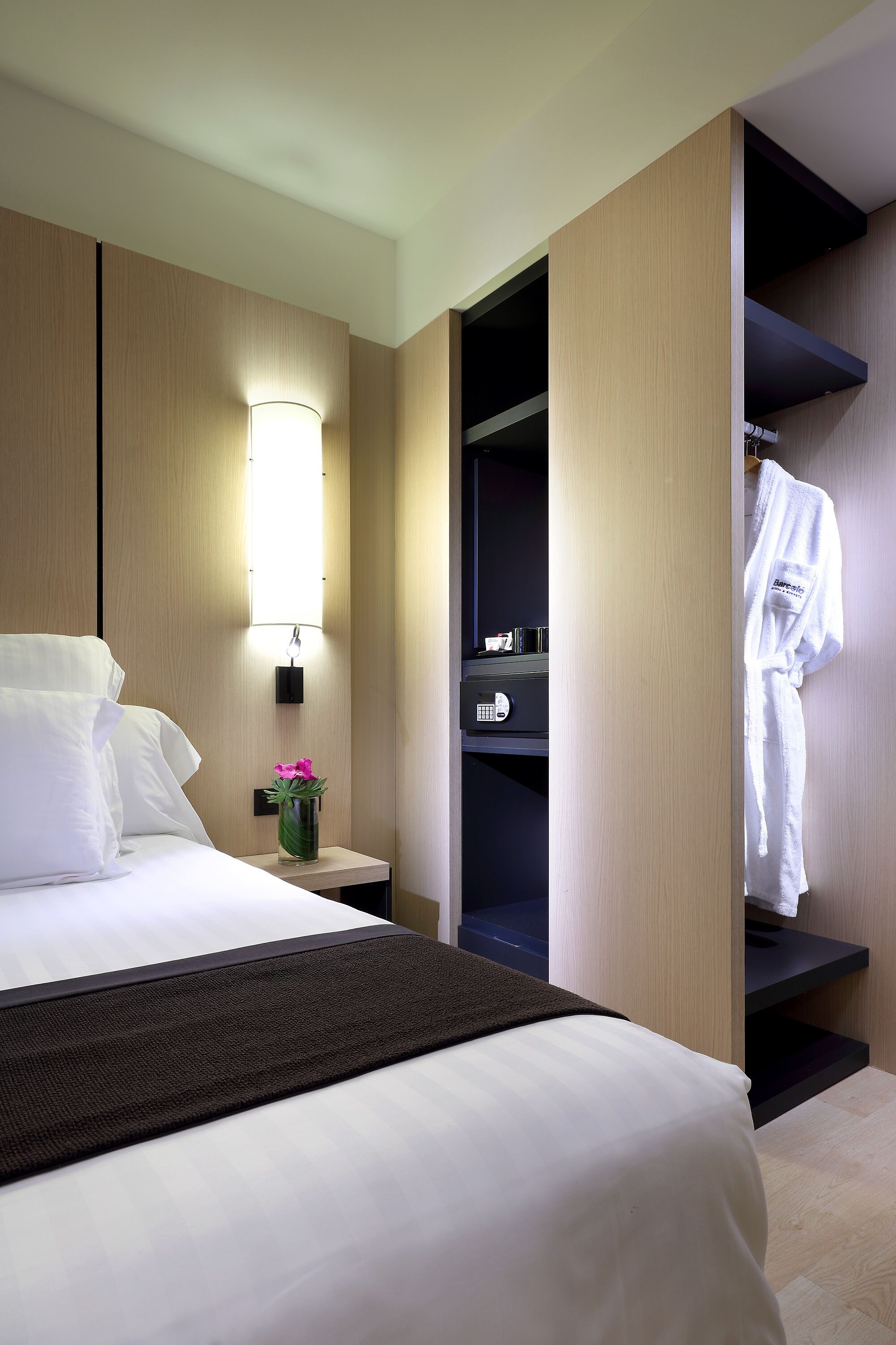 Una habitación de hotel con una cama, una mesita de noche con lámpara, un florero con flores, una bata colgada en un perchero y un armario con espejo.