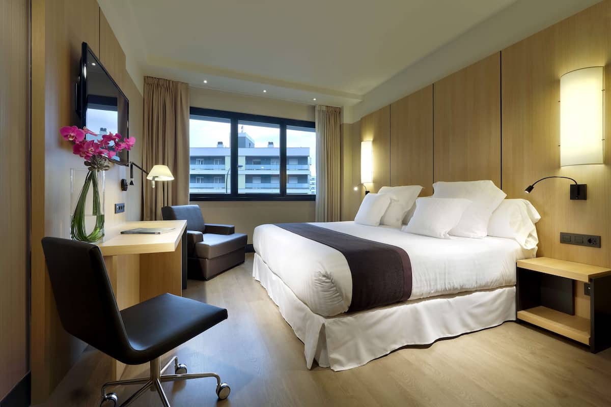 A modern hotel room with a large bed, a desk, a chair, and a TV mounted on the wall.