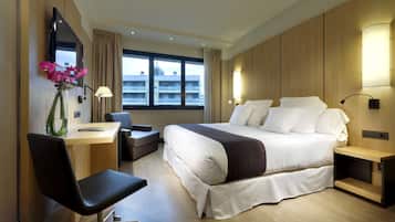 Quarto de hotel moderno com uma cama grande, escrivaninha, cadeira e televisão na parede.