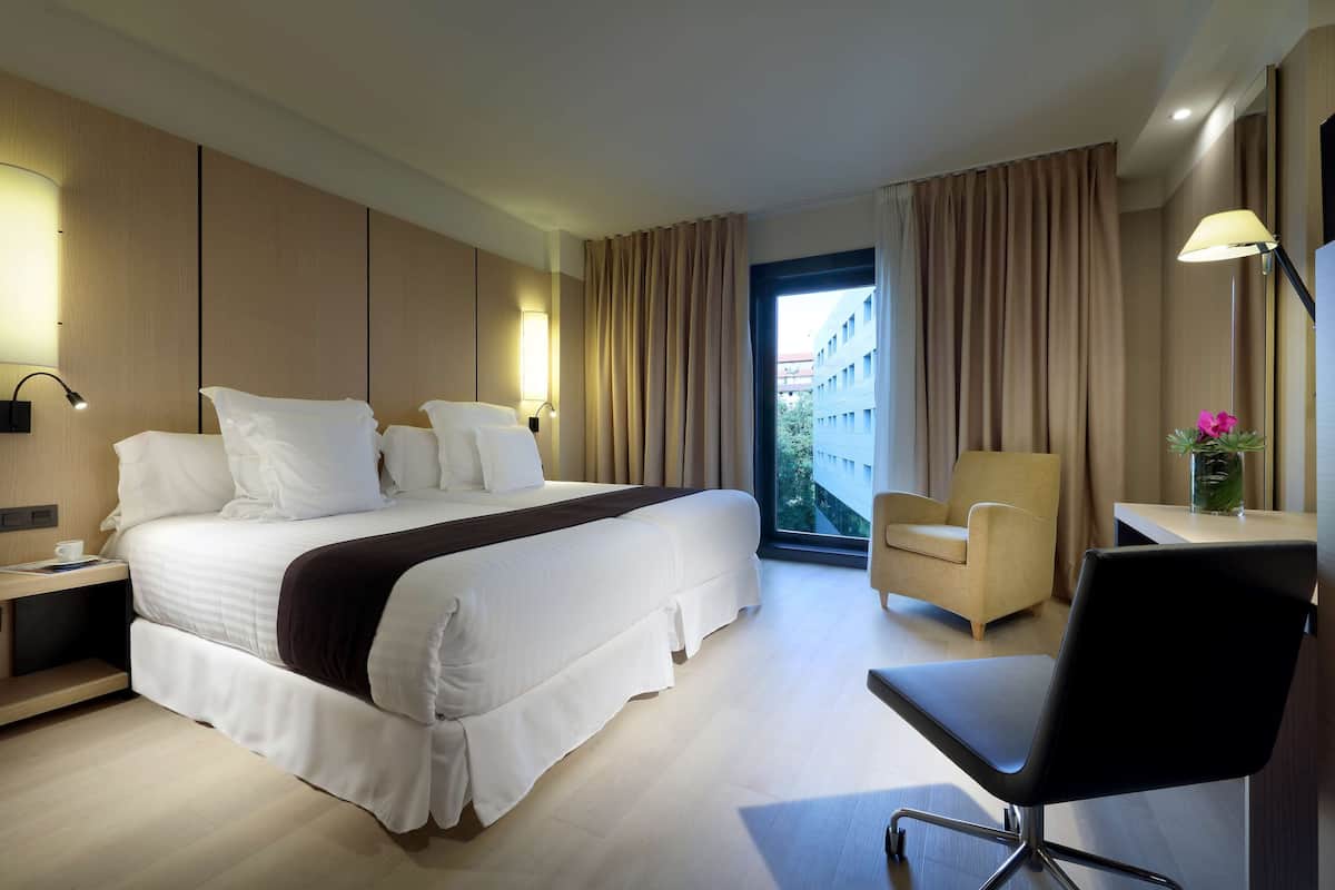 A modern hotel room with a large bed, a desk, a chair, and a view of the city through the window.
