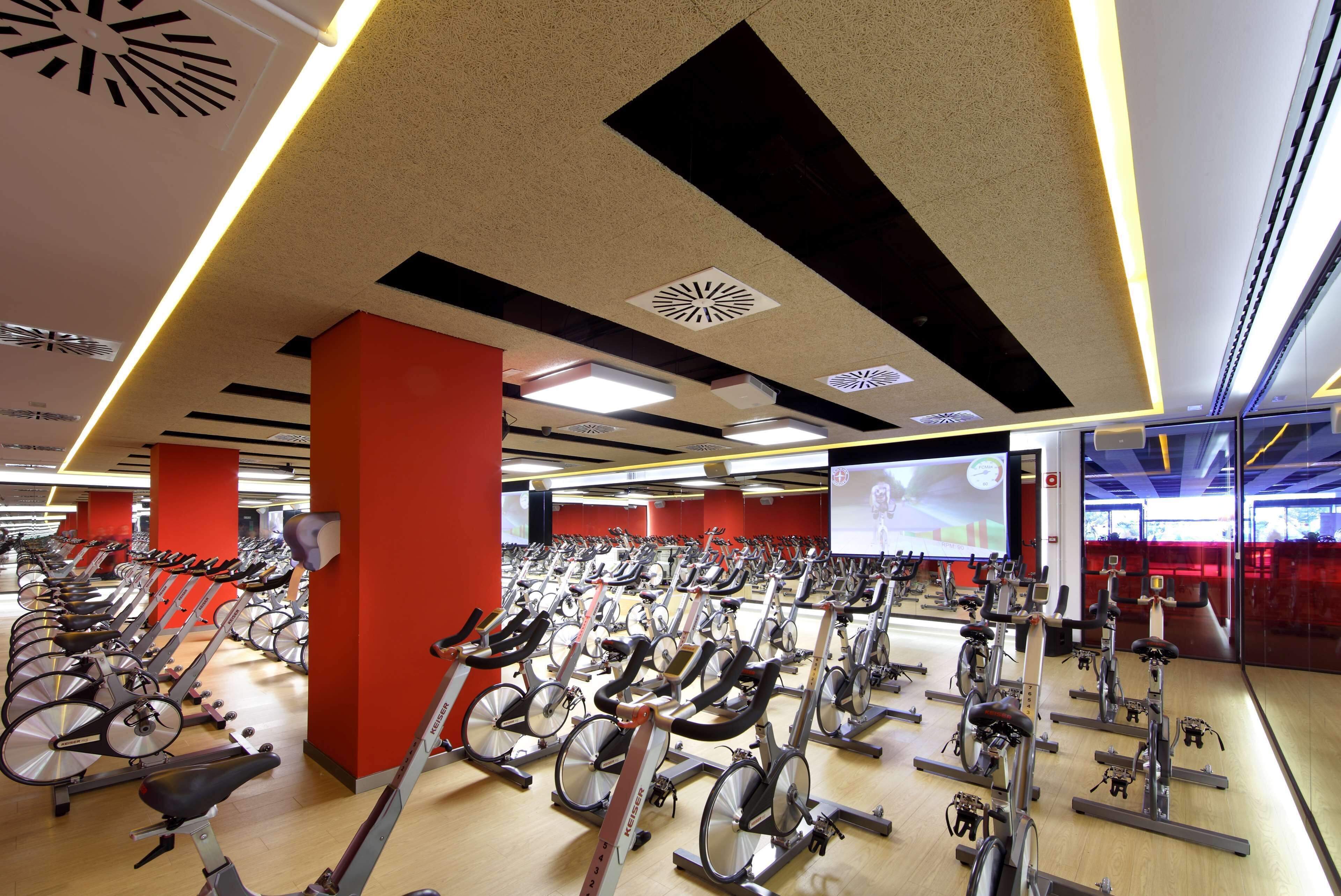 A gym with rows of stationary bikes, red pillars, and a large screen in the background.