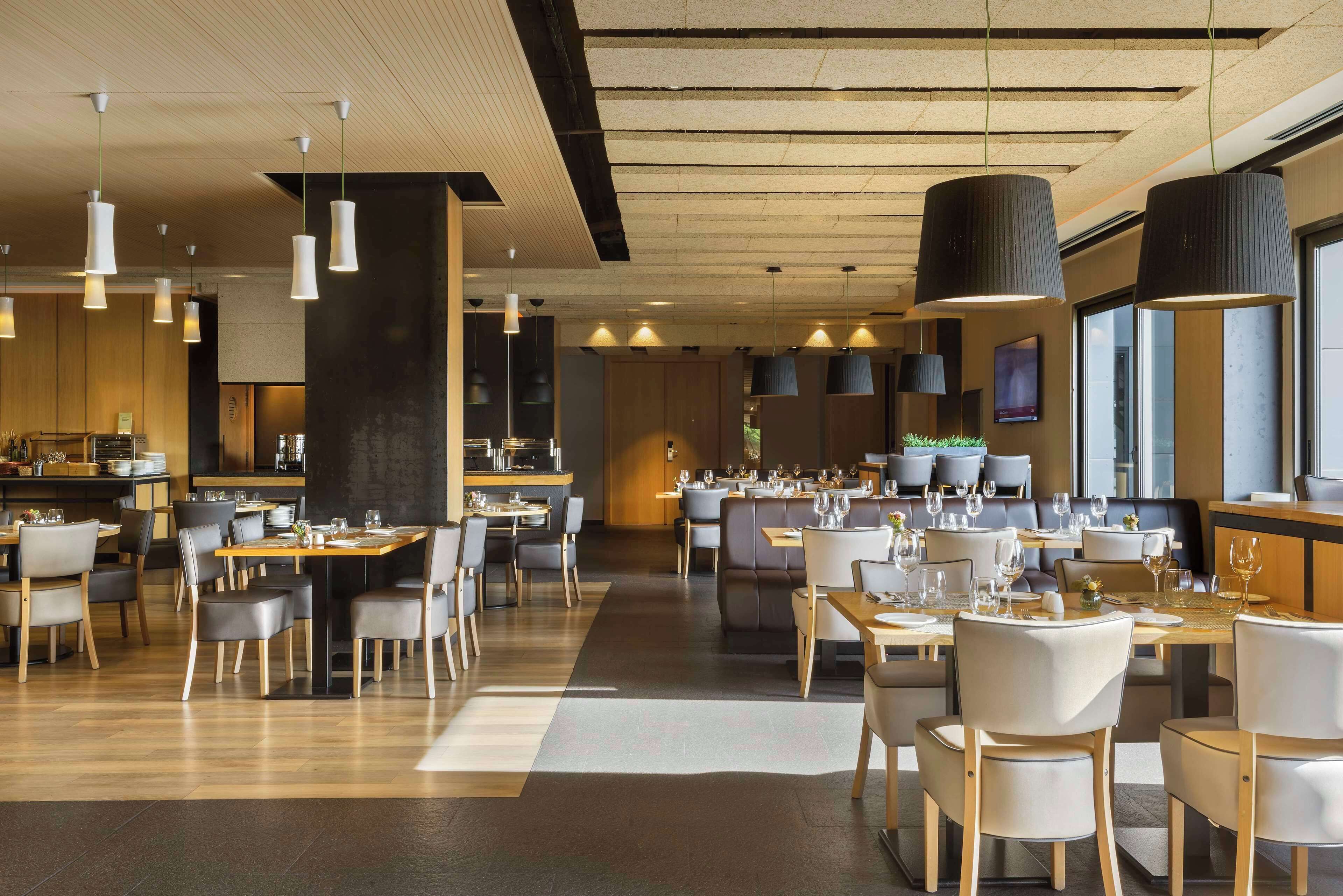 A modern restaurant with multiple dining areas, wooden tables, and contemporary lighting fixtures.