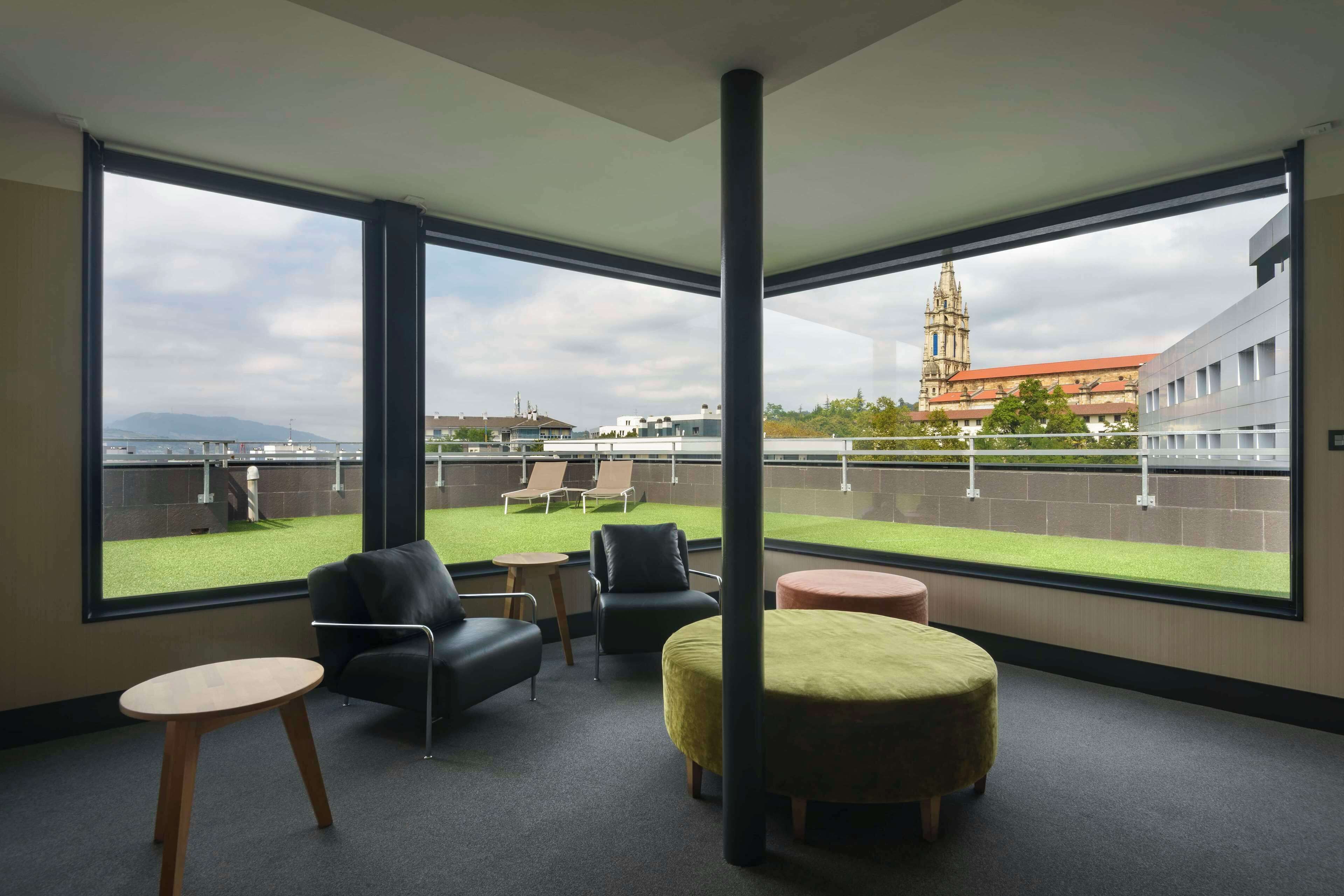 Une chambre moderne dotée de grandes fenêtres, offrant une vue sur un bâtiment historique, et disposant d’un petit espace extérieur aménagé pour s’asseoir.