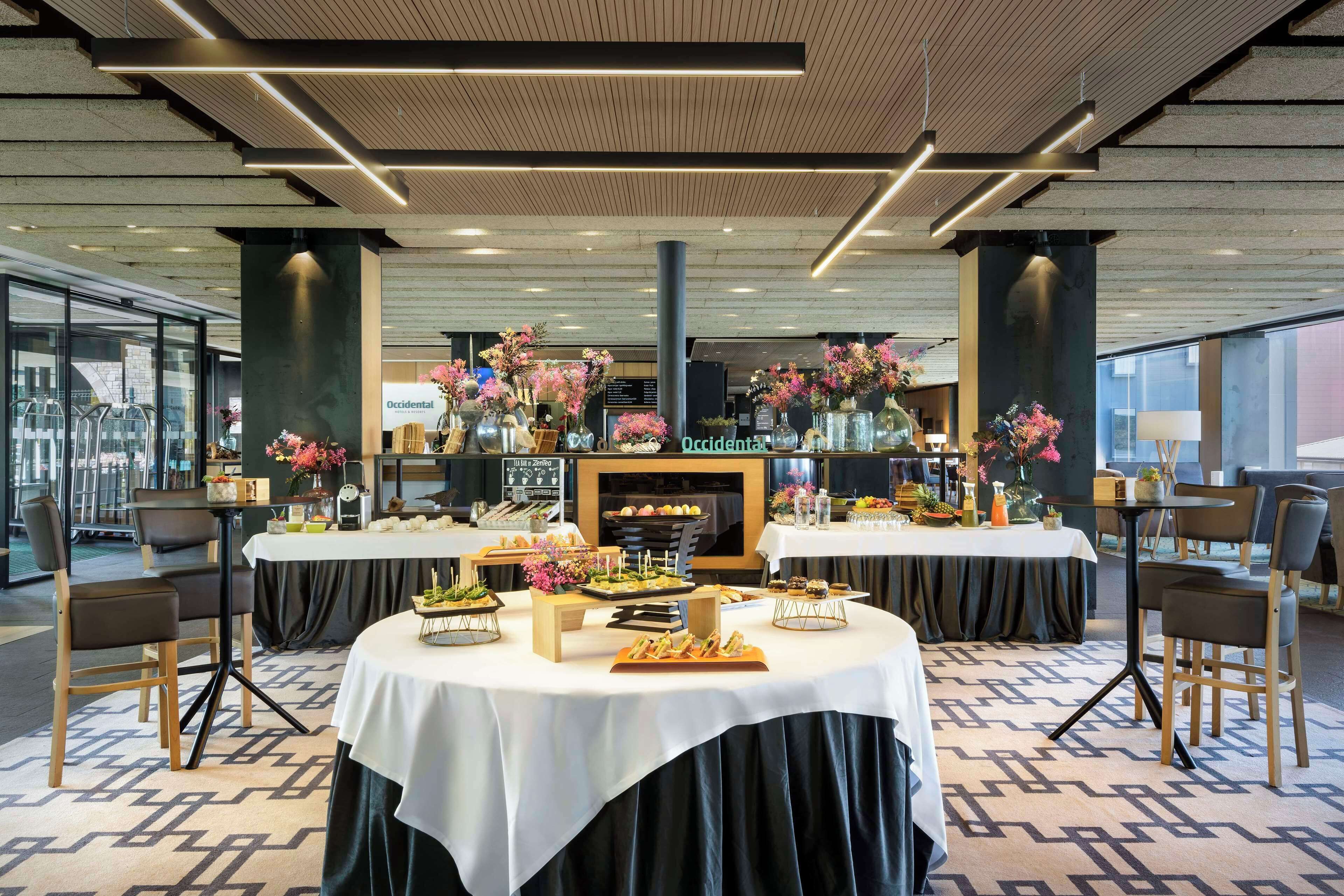 Eine moderne Hotelhalle mit einem zentralen Empfangsbereich, mehreren Tischen mit floralen Dekorationen und einem großen Bildschirm, der den Namen des Hotels zeigt.