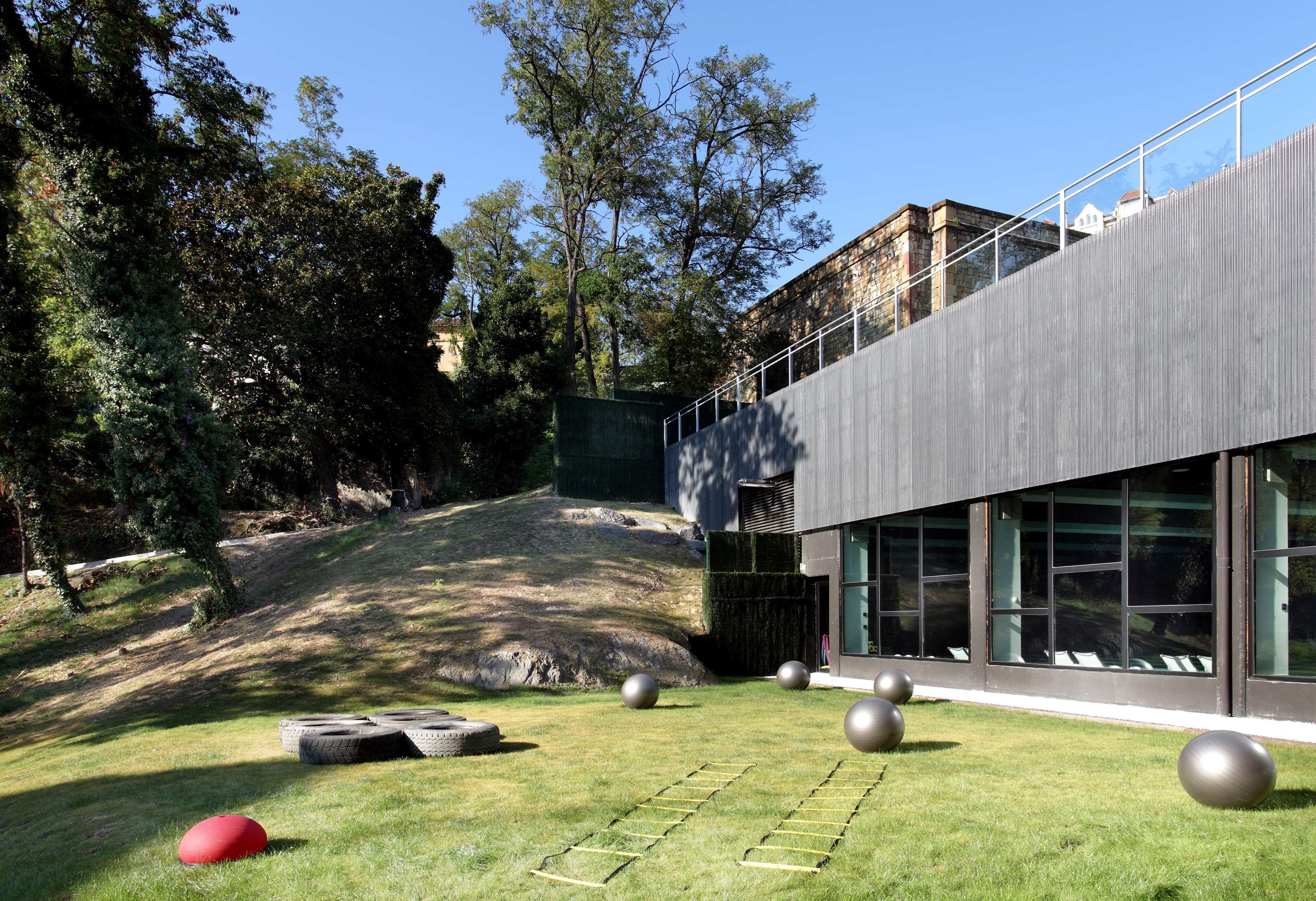 Un edificio moderno con grandes ventanales, un área verde con equipamiento de ejercicio y árboles al fondo.