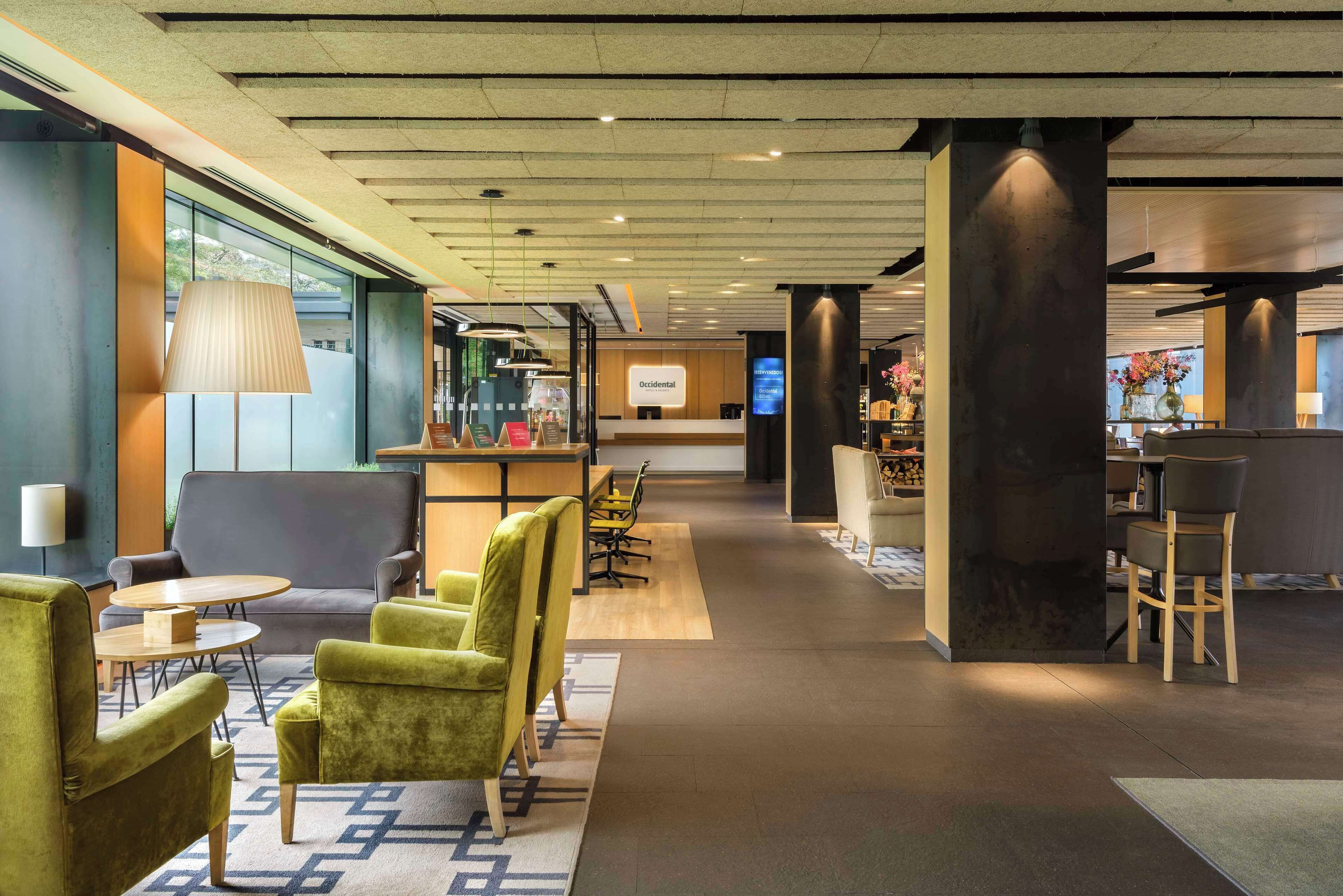 Un hall d’hôtel moderne avec des fauteuils verts, un comptoir d’accueil et un espace bar.