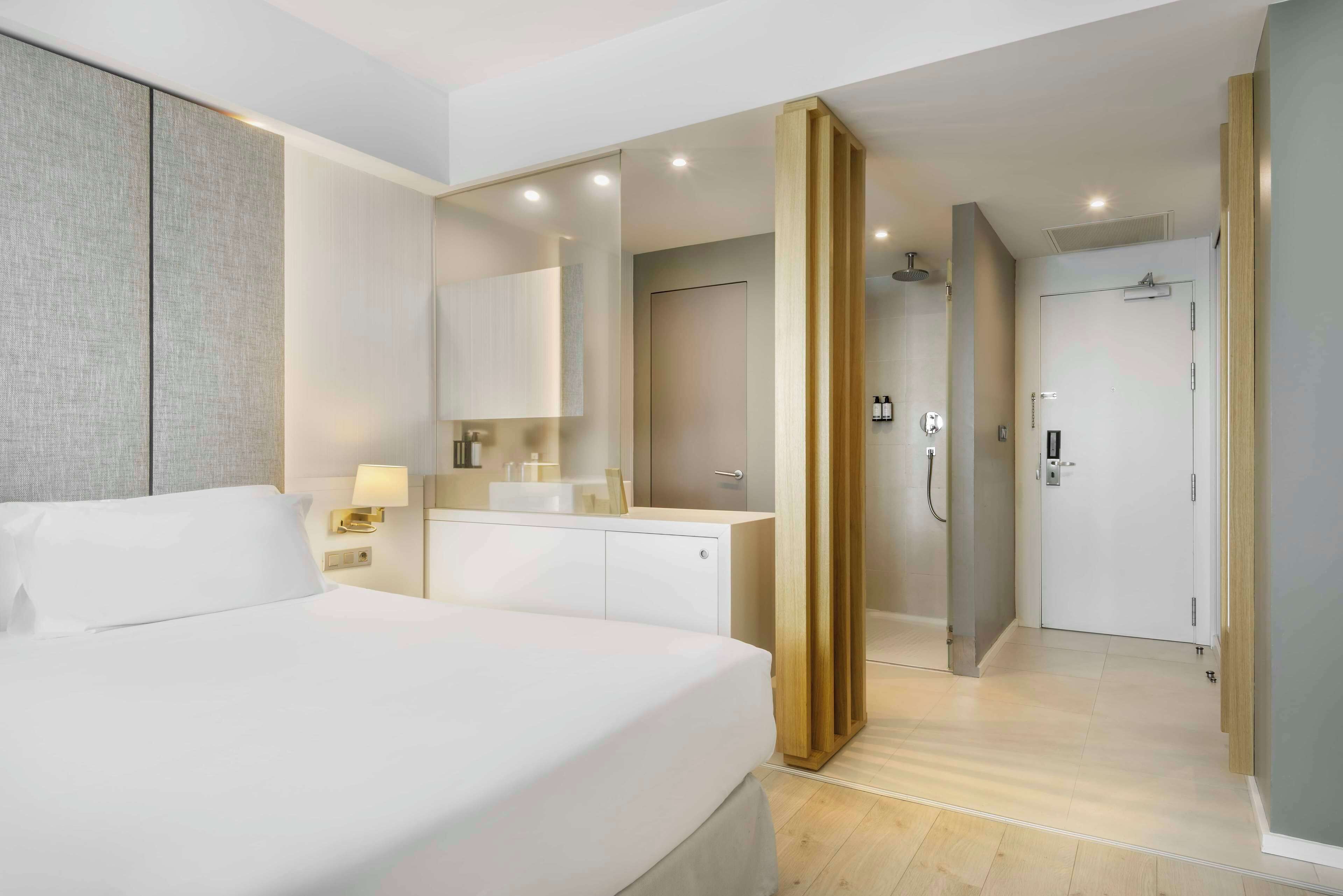 Habitación de hotel moderna con una cama grande, una mesita de noche, un espejo, un baño con ducha y una puerta que da a otra habitación.