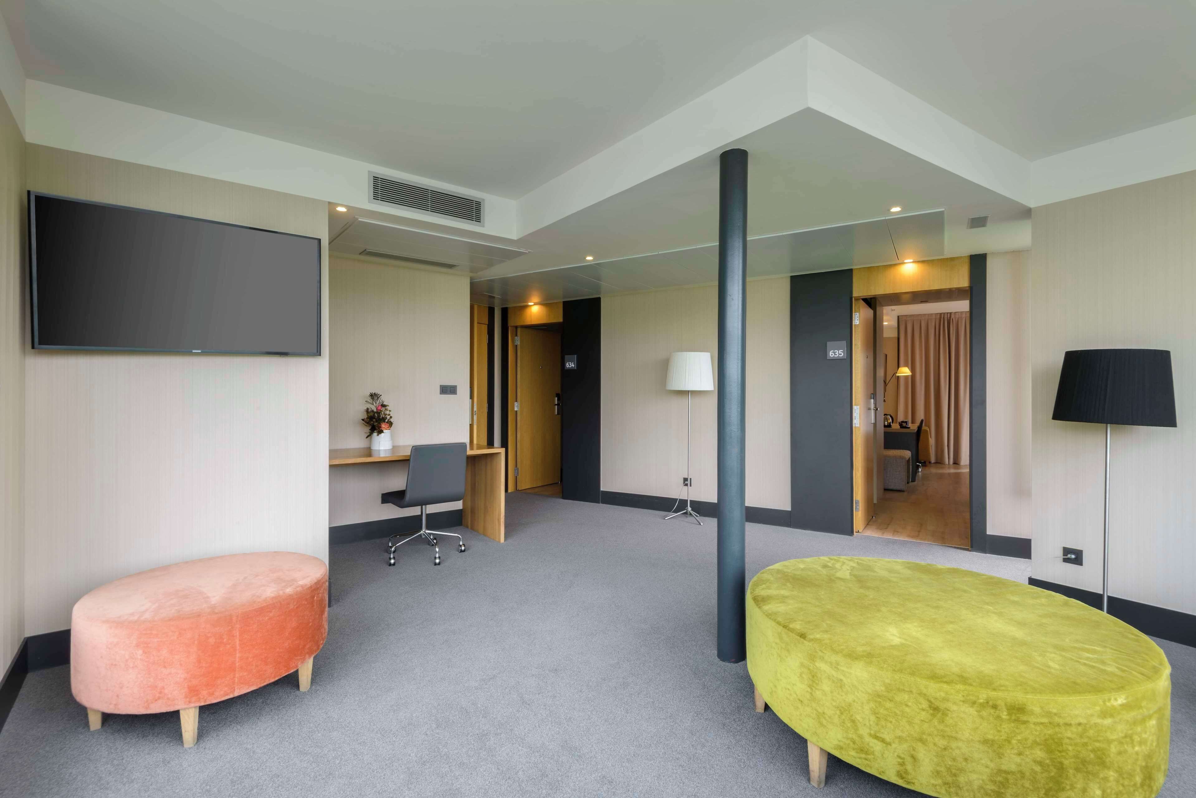 A modern hotel room with a large flat-screen TV, a sofa, a desk with a chair, and a lamp.