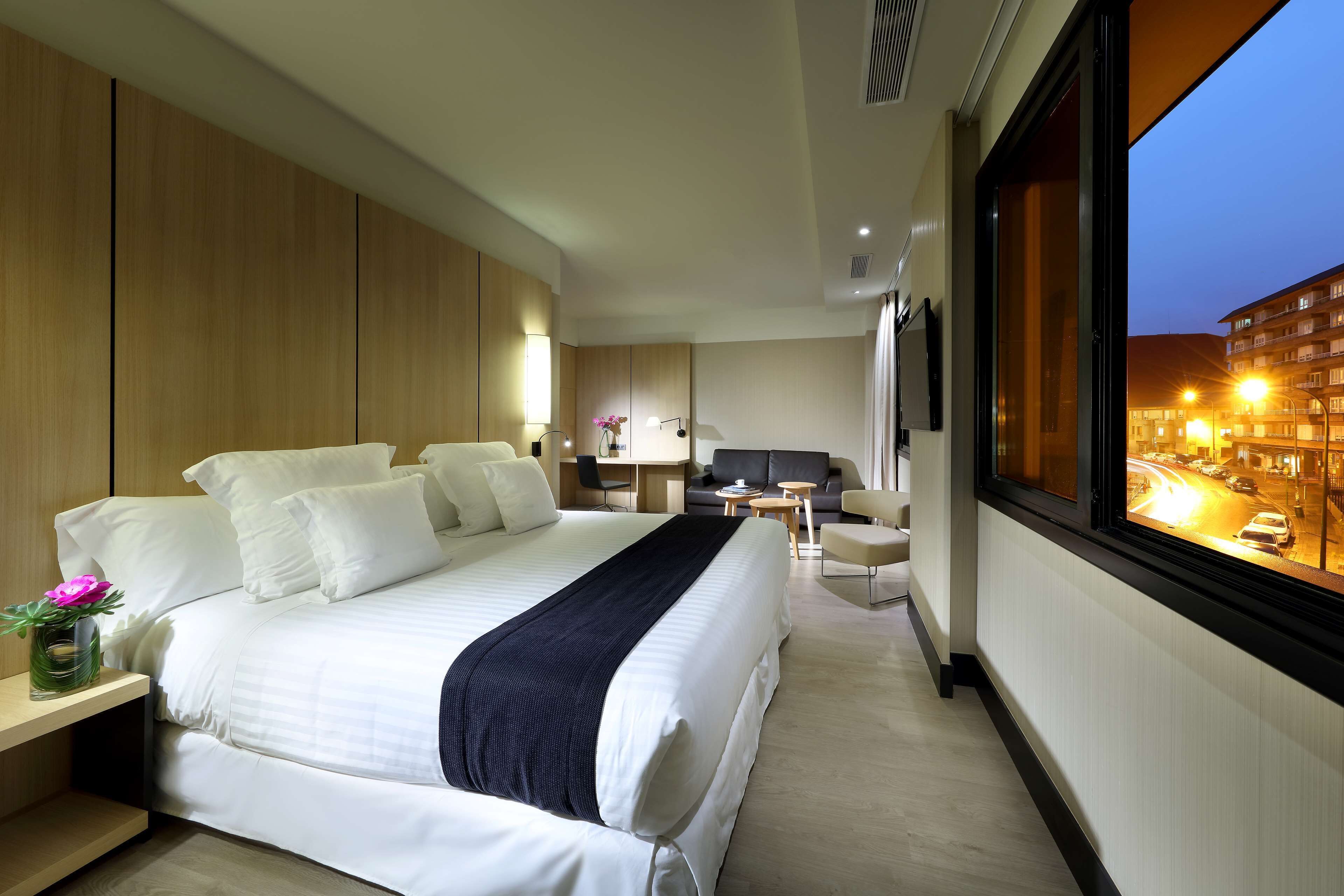Quarto de hotel moderno com uma cama grande, uma escrivaninha e vista para a paisagem urbana ao entardecer.