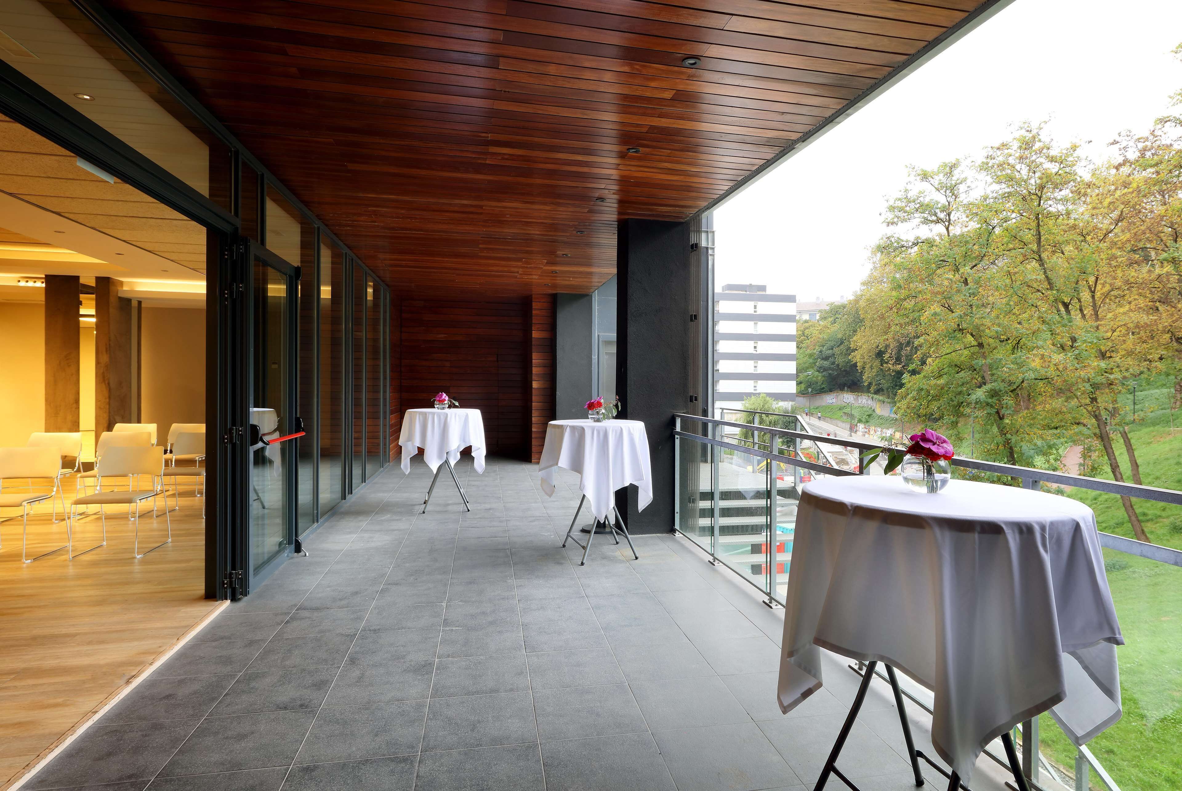 Un espacio al aire libre techado con mesas dispuestas para comer, techo de madera y vistas a árboles y edificios al fondo.