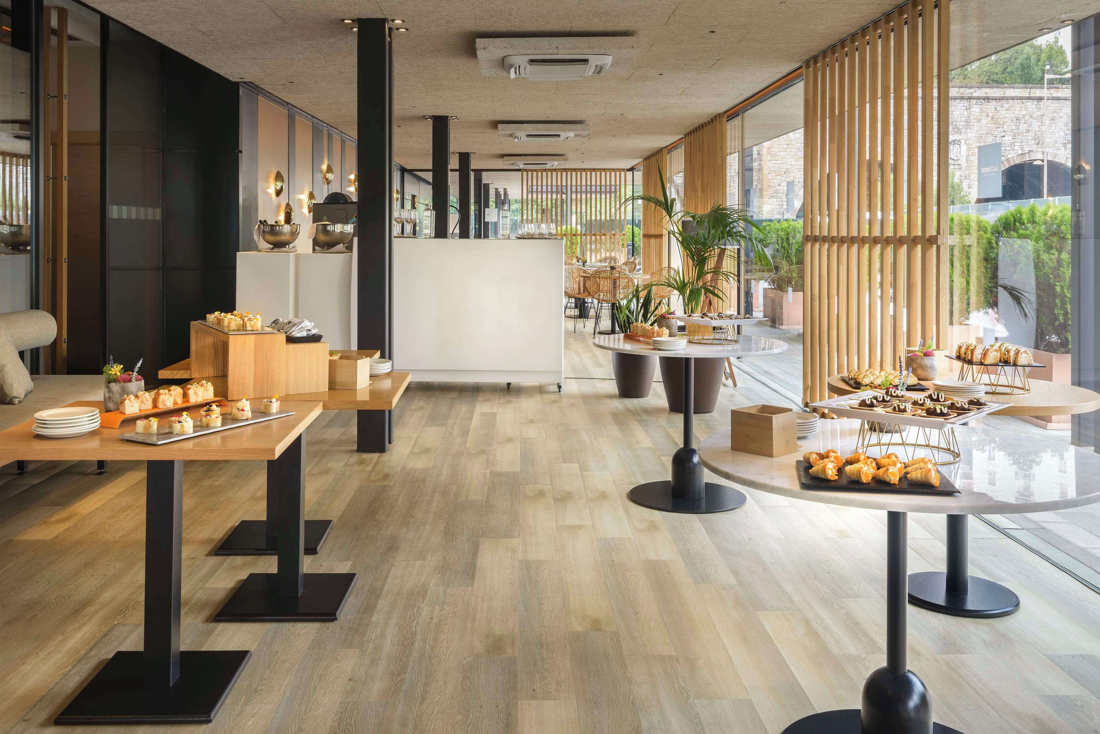 Un comedor moderno con mesas y sillas de madera, una isla central con bufé, y grandes ventanales que ofrecen vistas al exterior.
