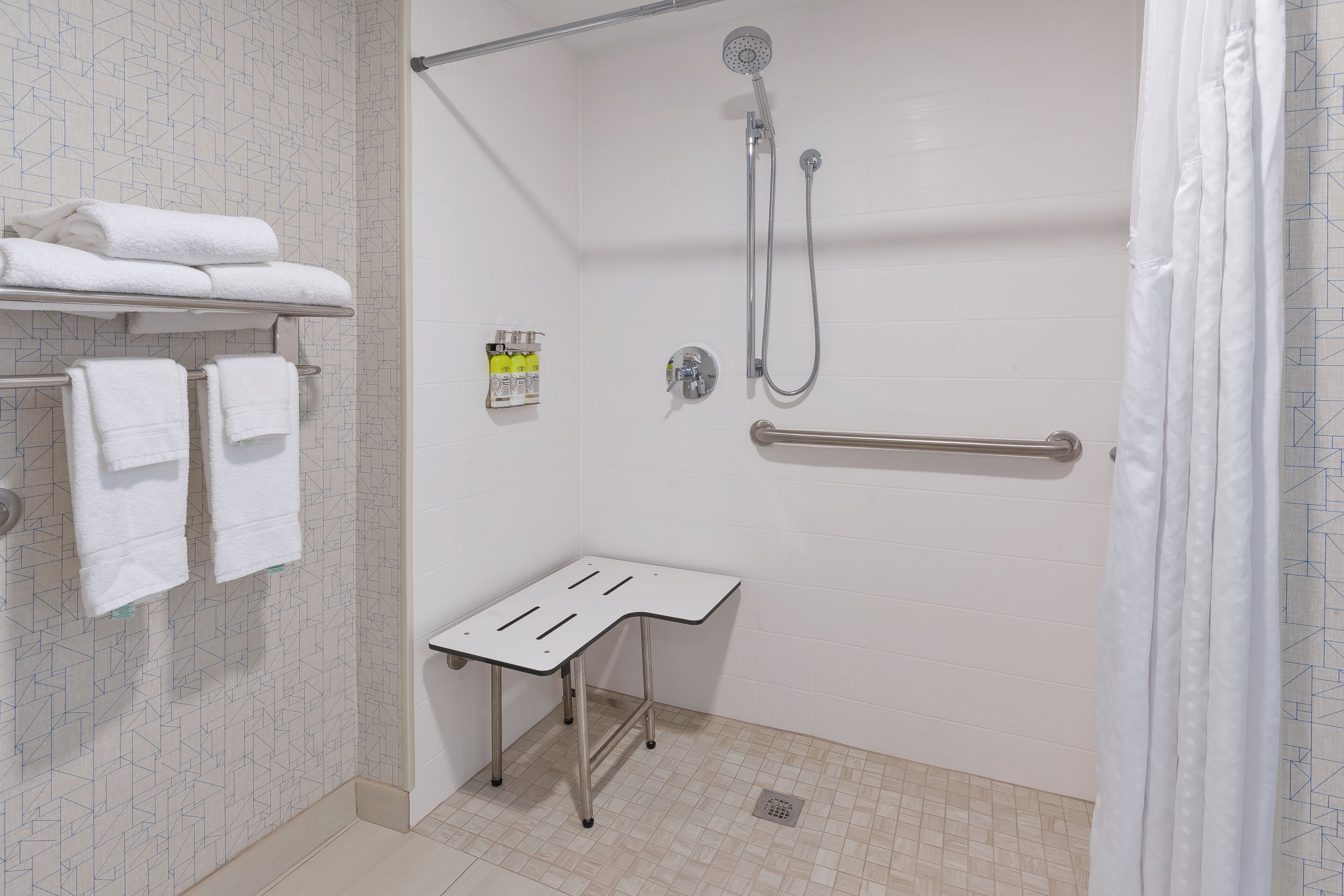 Un espace douche équipé d’un pommeau de douche à main, d’un porte-serviettes avec des serviettes pliées, d’une petite table et d’une barre d’appui.