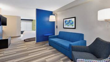 A hotel room with a blue accent wall, a grey sofa, a patterned armchair, a flat-screen TV, and a framed abstract artwork.