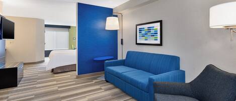 A hotel room with a blue accent wall, a grey sofa, a patterned armchair, a flat-screen TV, and a framed abstract artwork.
