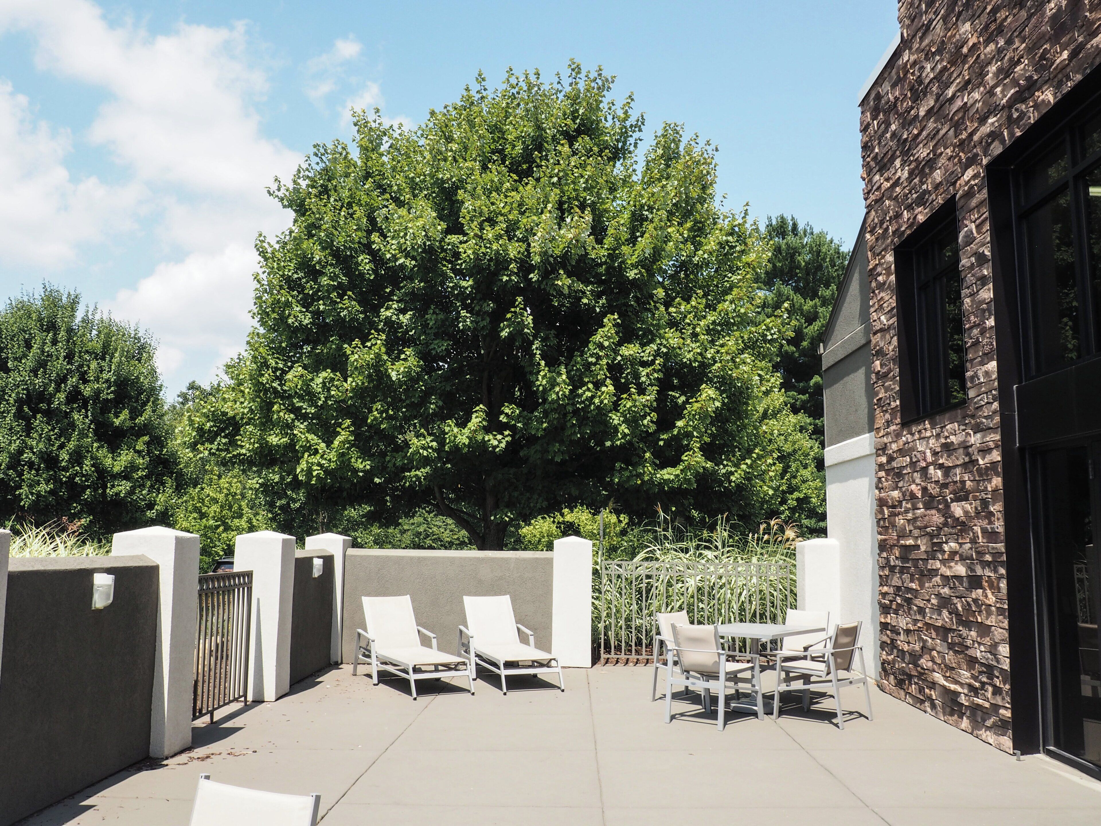 An outdoor patio with white lounge chairs and a table, a stone wall, and a large tree in the background.