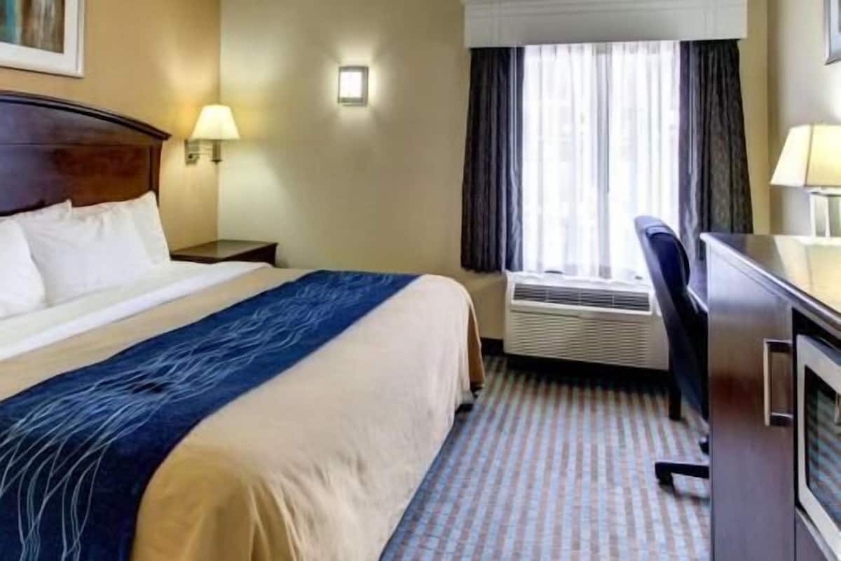 Ein Hotelzimmer mit einem Bett, einem Schreibtisch, einem Stuhl, einem Fenster mit Vorhängen, einer Klimaanlage und einer Wandlampe.