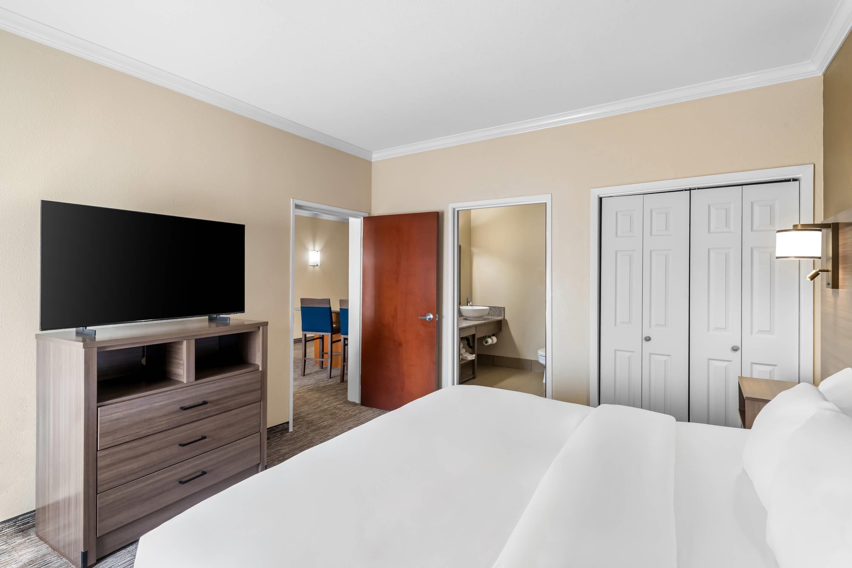 Quarto de hotel com cama, televisão sobre suporte de madeira, guarda-roupa e casa de banho visível através de uma porta aberta.