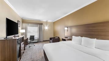 A hotel room with a large bed, a desk with a flat-screen TV, a chair, a nightstand, and a window with curtains.