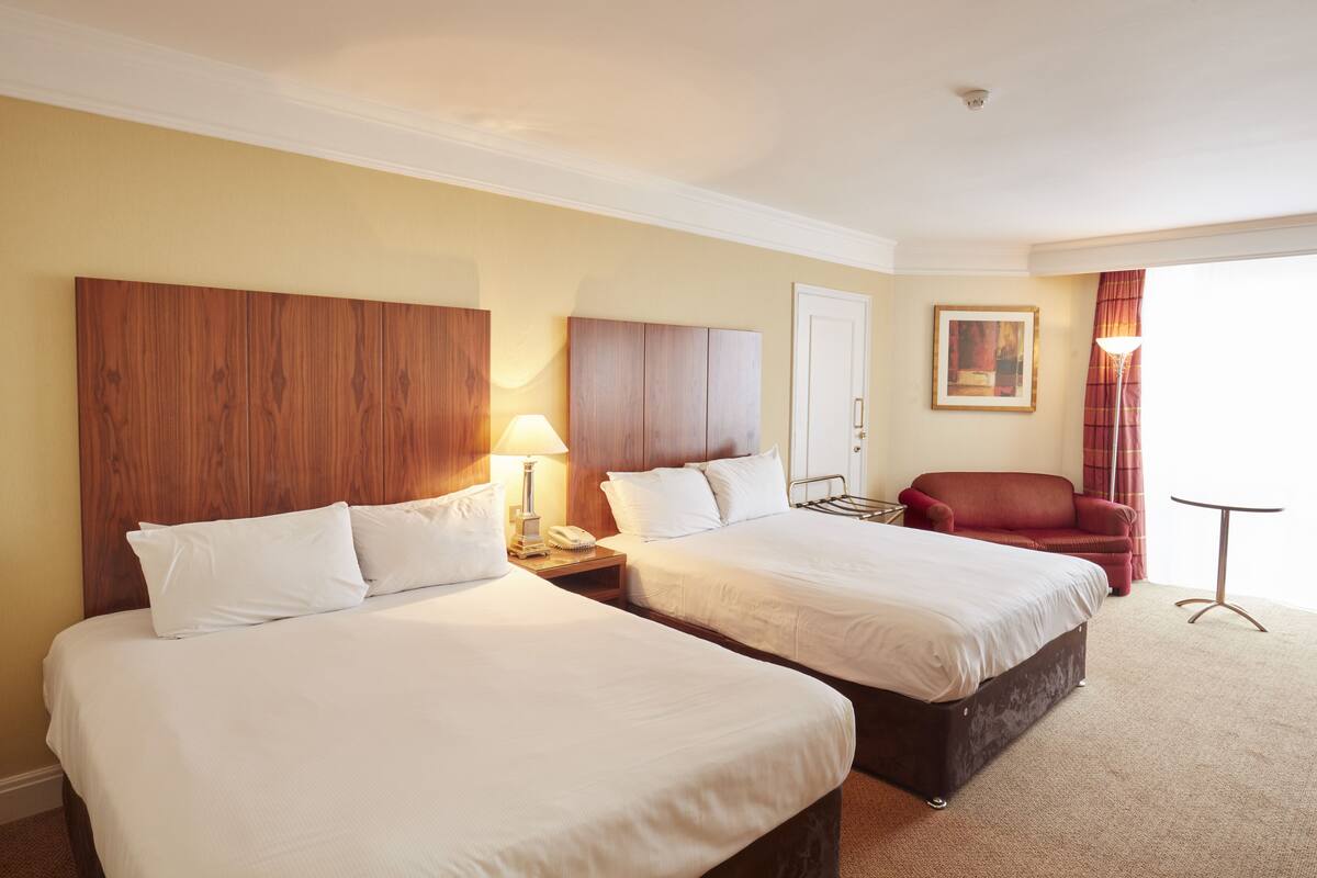 A hotel room with two beds, a wooden headboard, a red armchair, a small table, and a painting on the wall.