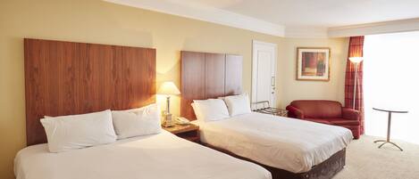 A hotel room with two beds, a wooden headboard, a red armchair, a small table, and a painting on the wall.