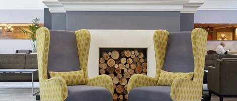 A lobby with a fireplace, two patterned armchairs, a sofa, and a stack of logs.