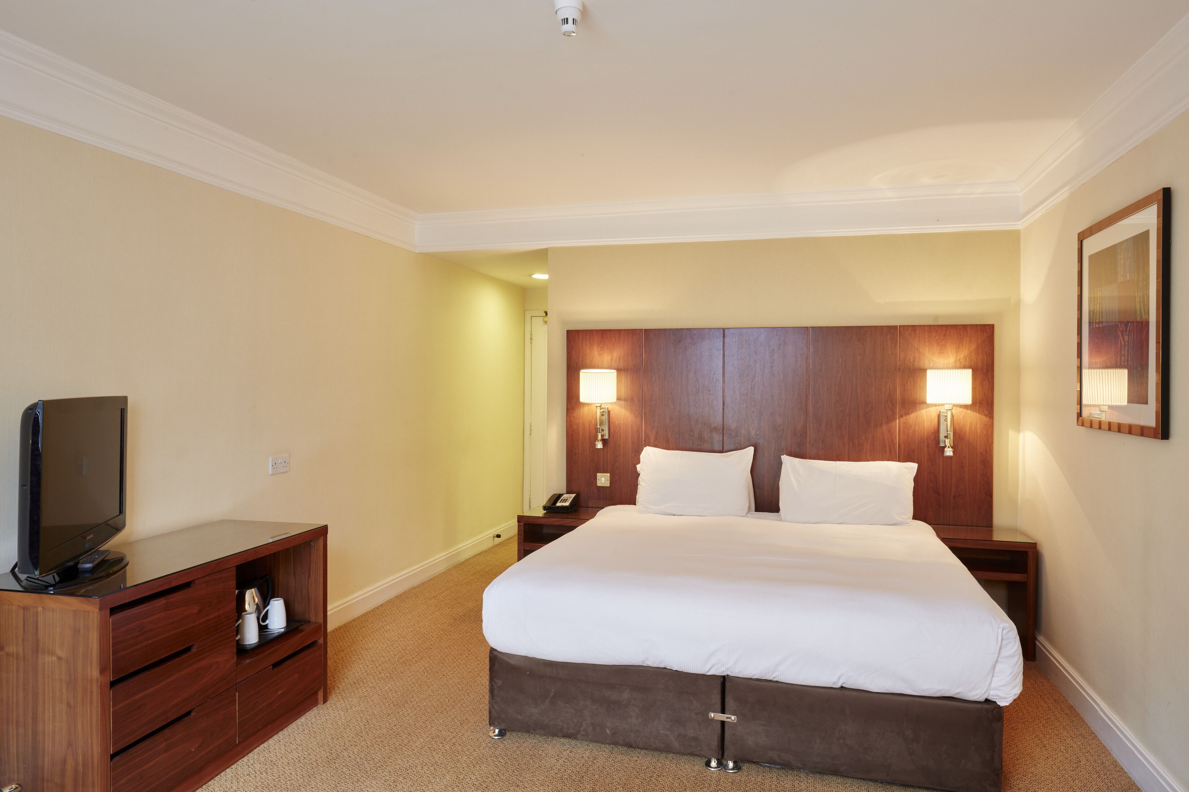A hotel room with a large bed, wooden headboard, bedside lamps, a TV on a wooden cabinet, and a small nightstand.