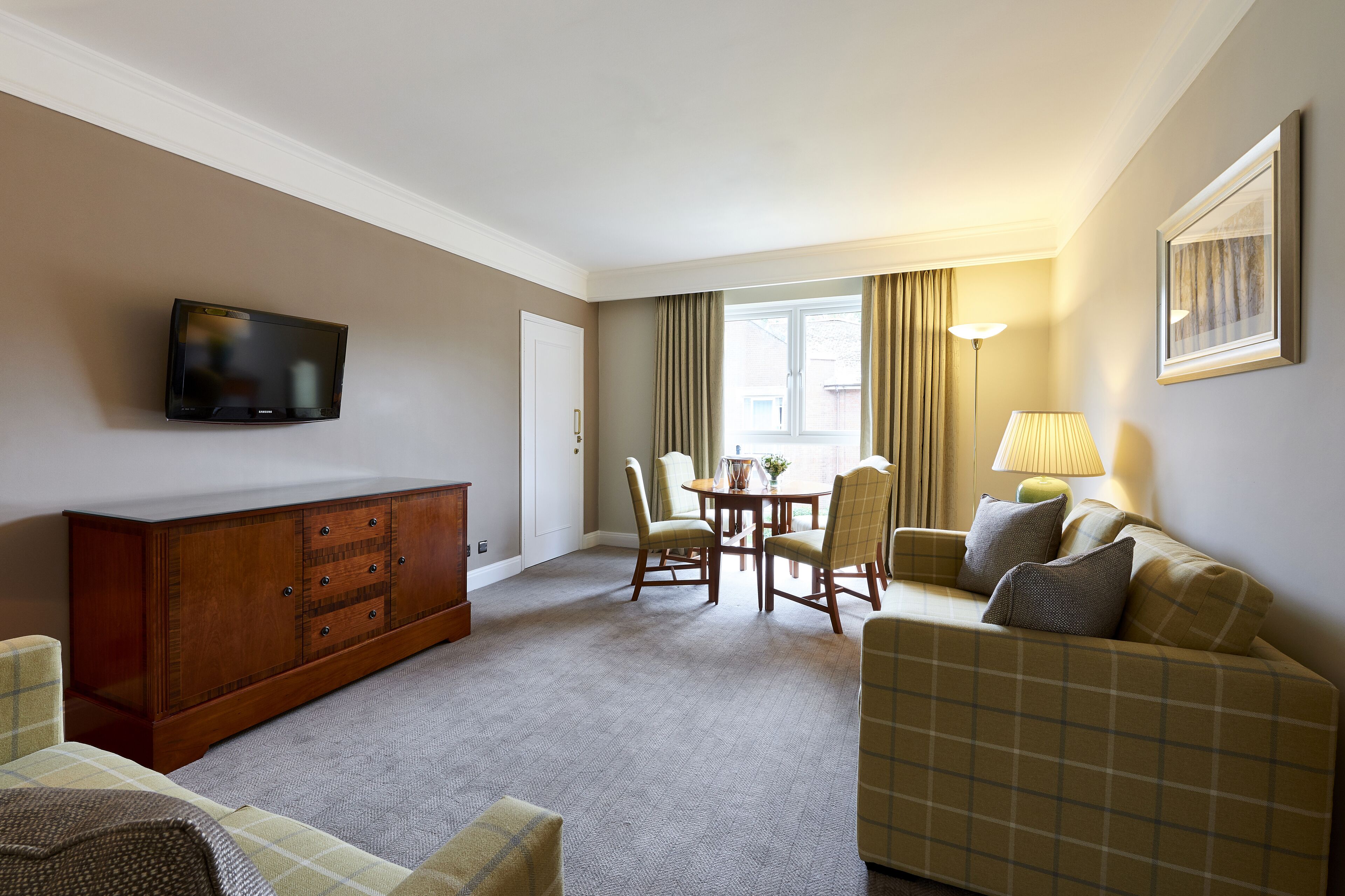 Habitación de hotel con televisión de pantalla plana, un armario de madera, una mesa de comedor con sillas, un sofá y una lámpara.