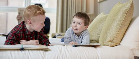 Dos niños leyendo en una cama con vista a la ventana y una lámpara al fondo.