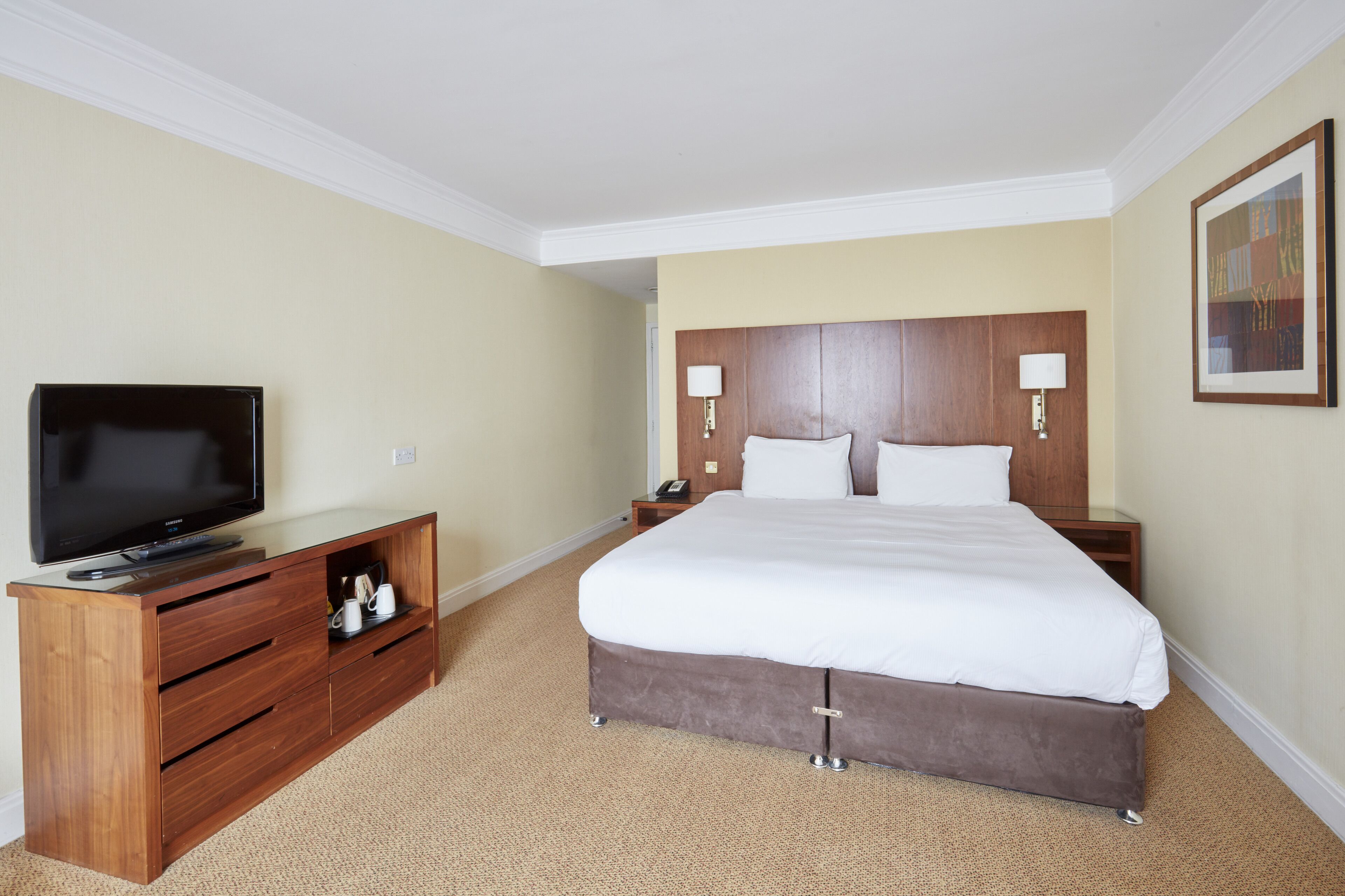 A hotel room with a large bed, a TV on a wooden console, and a nightstand with a lamp.