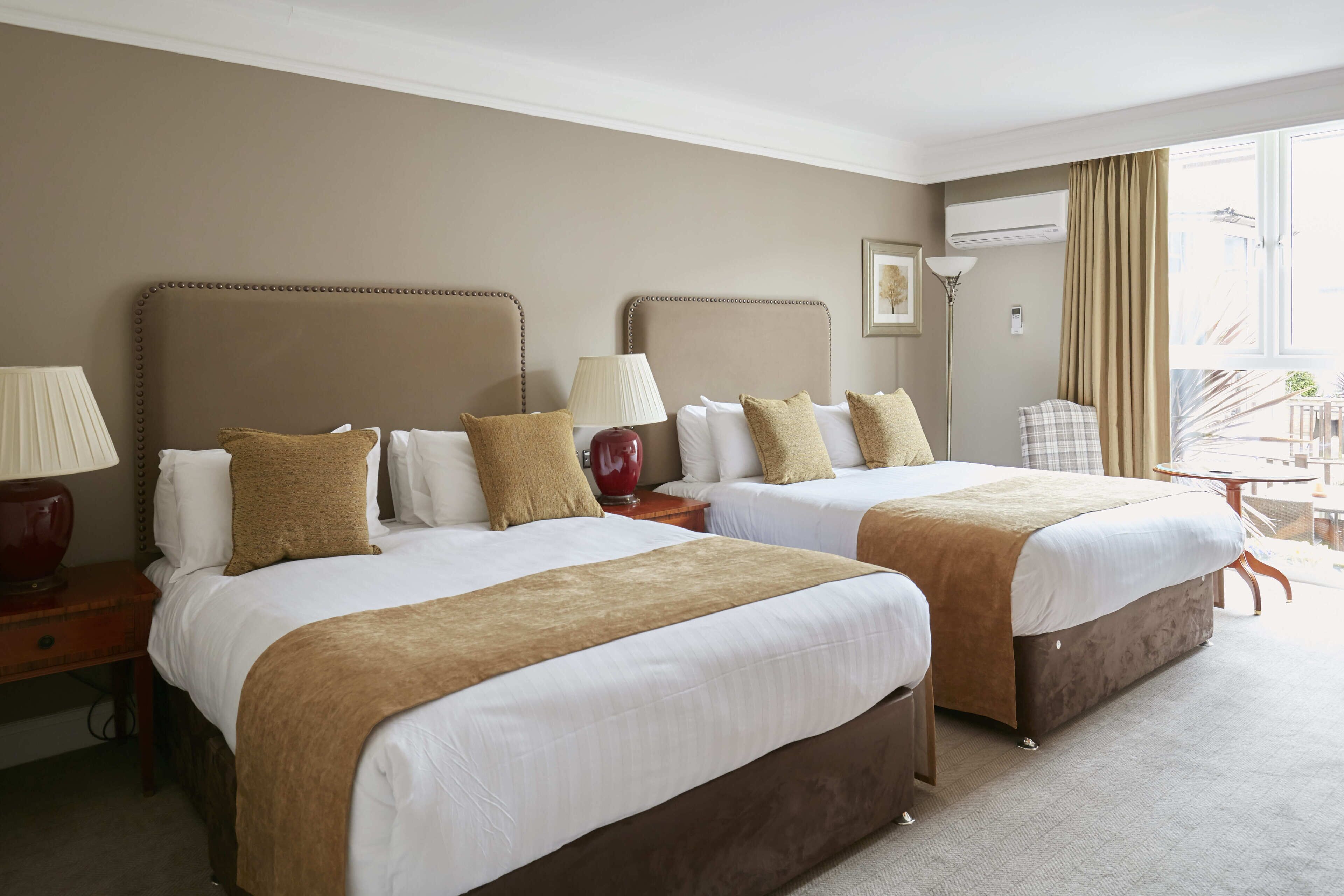 A hotel room with two beds, a lamp, a vase, and a view of the outside.