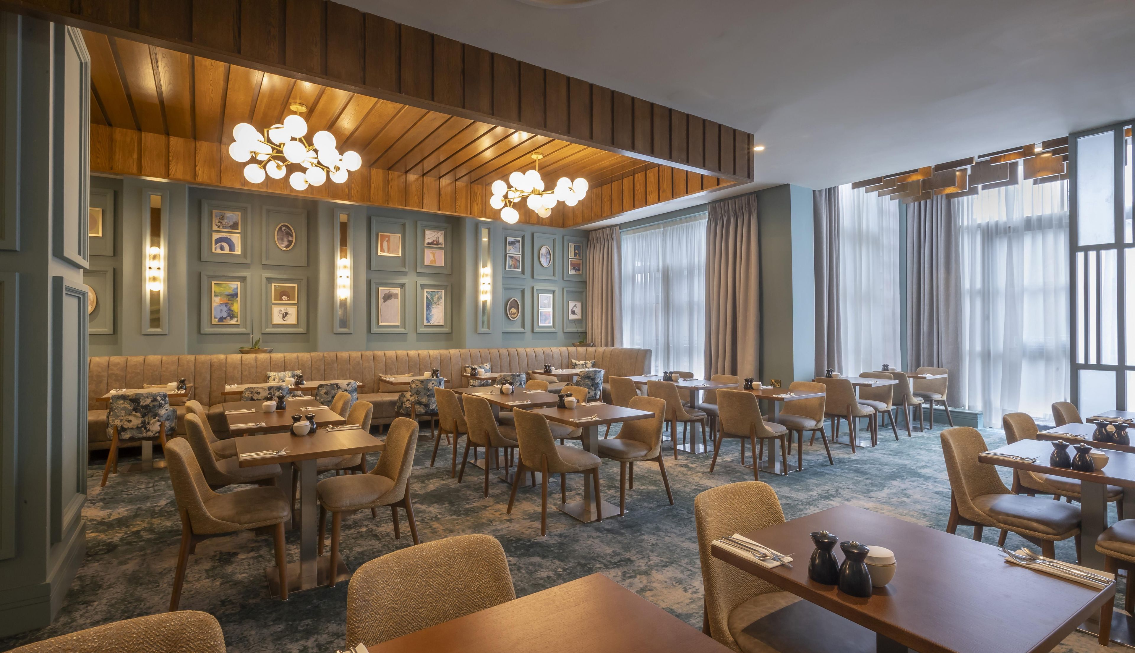 A hotel dining area with multiple tables and chairs, a high ceiling with wooden paneling, and wall-mounted picture frames.