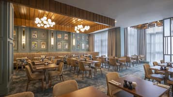 A hotel dining area with multiple tables and chairs, a high ceiling with wooden paneling, and wall-mounted picture frames.