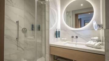 A modern bathroom with a glass-enclosed shower, a marble-tiled wall, a large mirror with a circular light, and a wooden vanity with a sink and towel rack.
