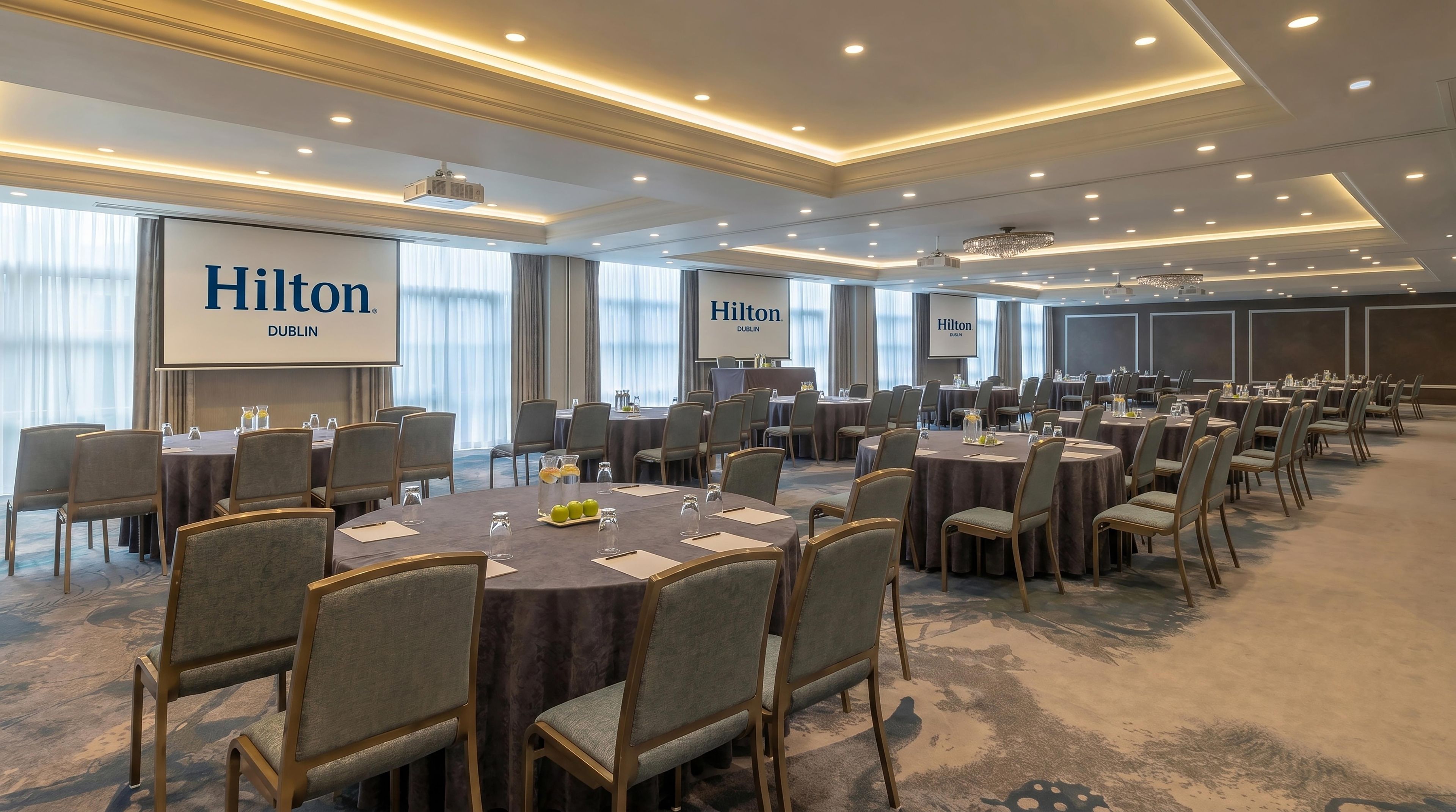Ein Konferenzraum mit mehreren Tischen und Stühlen, großen Fenstern und einer Projektionsleinwand, die 'Hilton Dublin' zeigt.