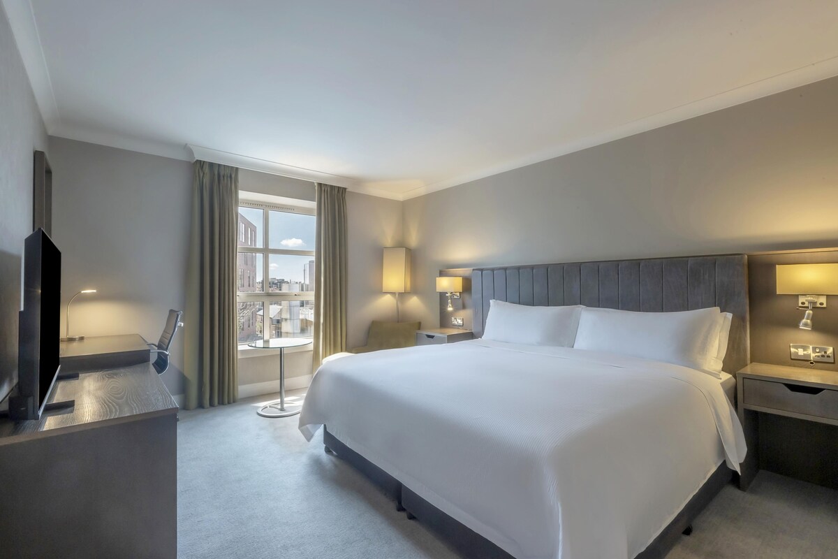 Une chambre d’hôtel avec un grand lit, un bureau, une chaise, une télévision et une fenêtre avec des rideaux.