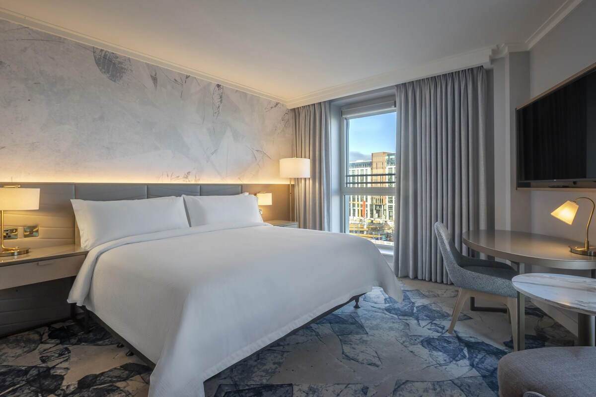 Une chambre d’hôtel avec un grand lit, un bureau avec une chaise, une télévision et une vue sur les immeubles par la fenêtre.