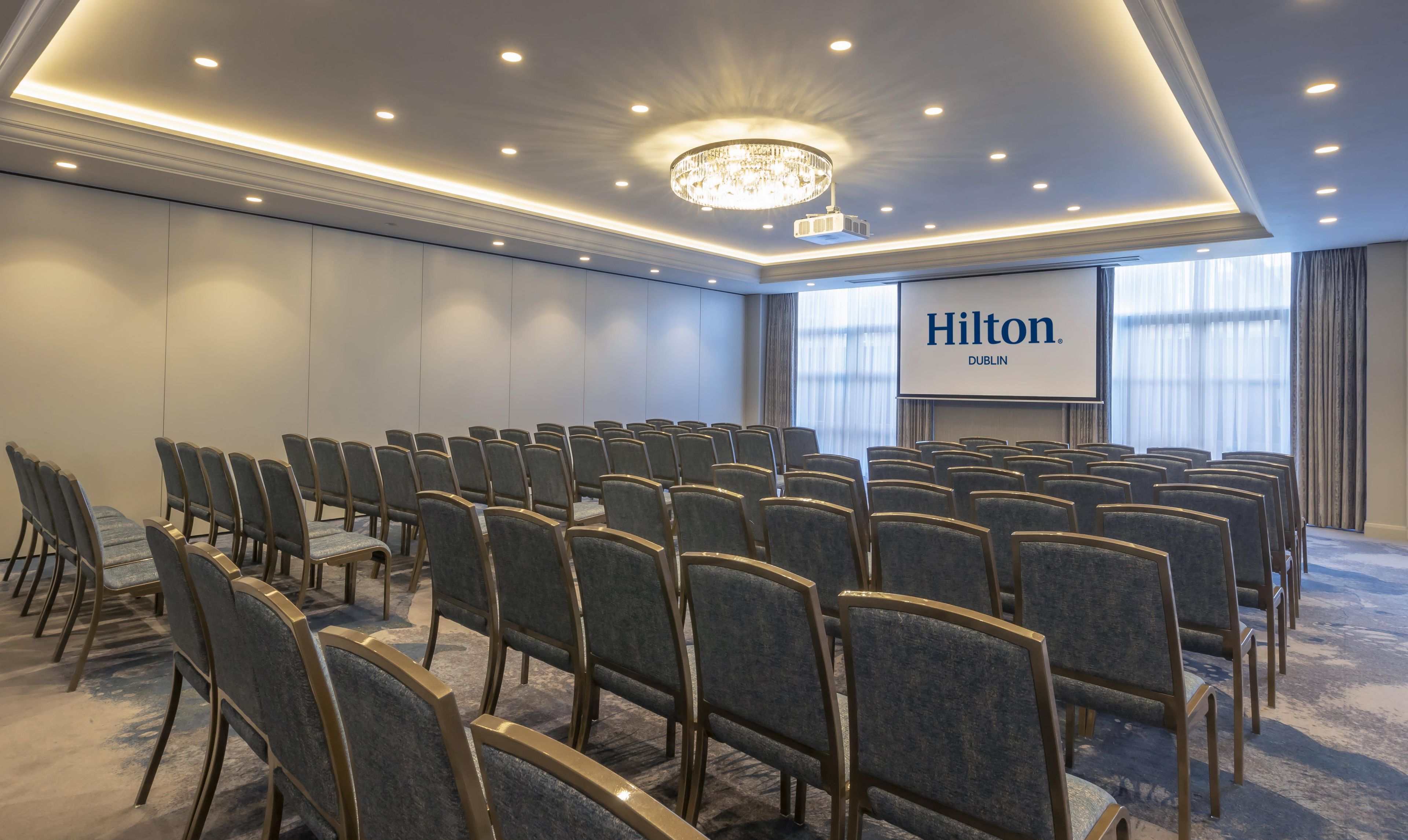 Et mødelokale med rækker af stole, en lysekrone og en skærm, der viser 'Hilton Dublin'.