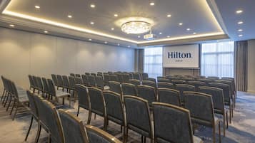 Et mødelokale med rækker af stole, en lysekrone og en skærm, der viser 'Hilton Dublin'.