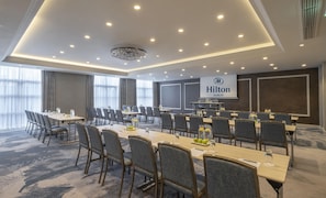 Une salle de conférence dotée de plusieurs tables et chaises, d’un grand écran affichant le logo de l’hôtel Hilton de Dublin, et d’un lustre.