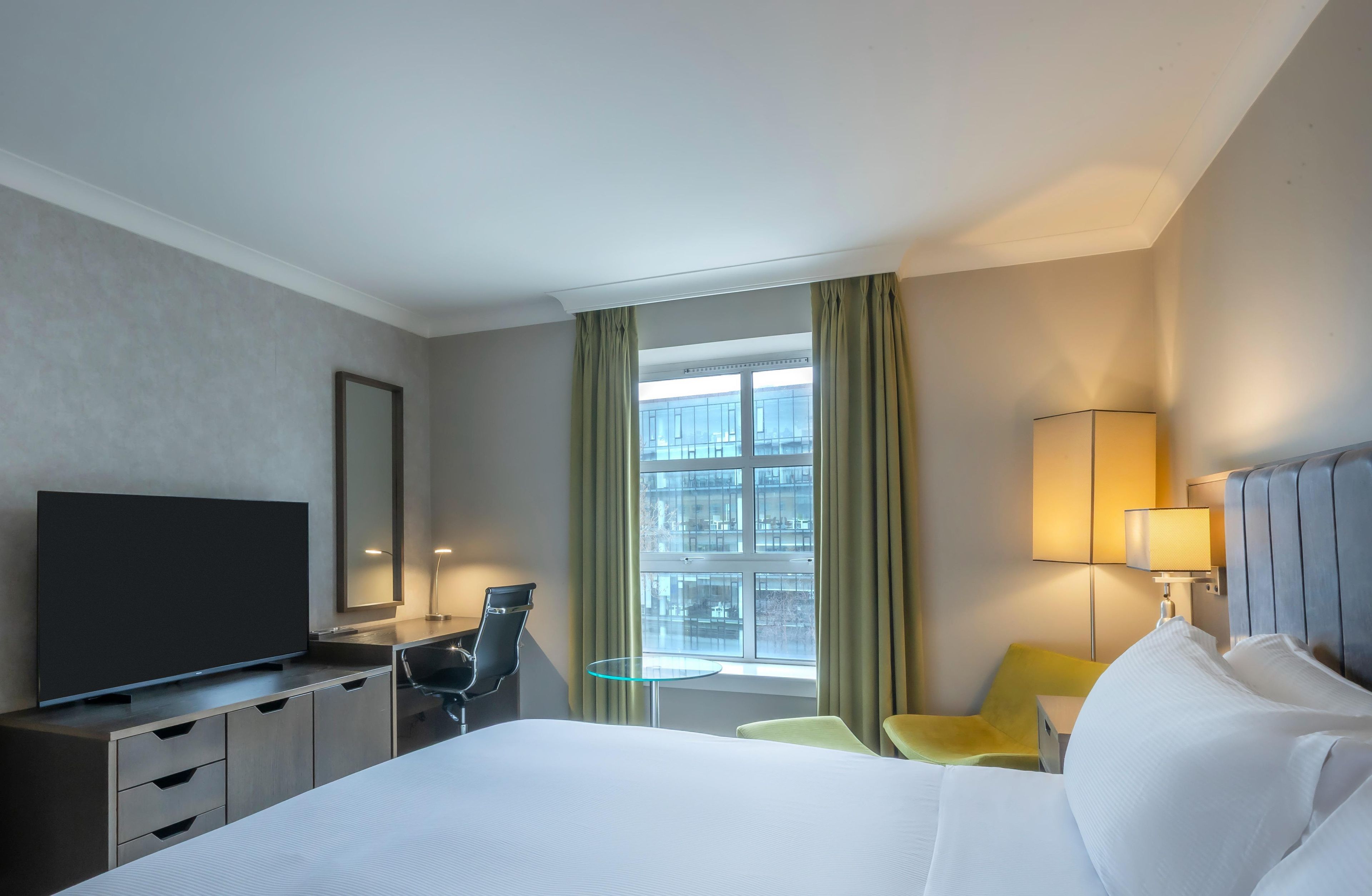 A hotel room with a bed, a desk, a television, a chair, a lamp, and a window with curtains.